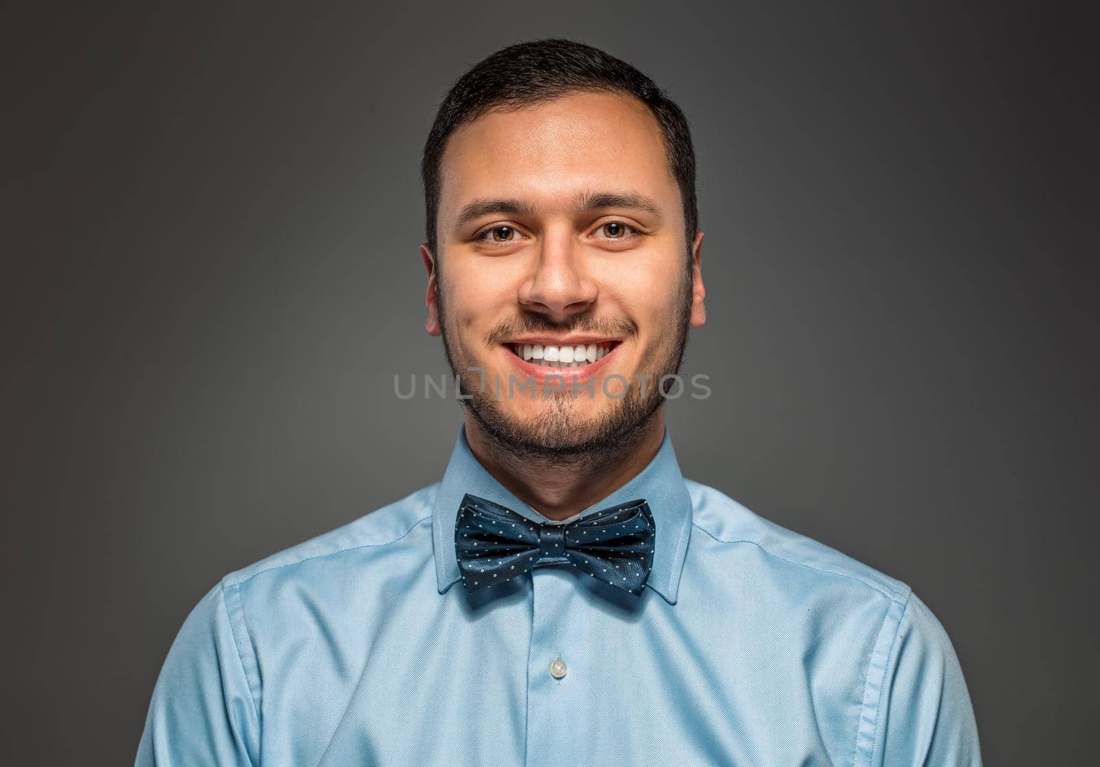 Portrait of happy smiling young man in blue shirt and butterfly tie isolated over gray studio background. Closeup