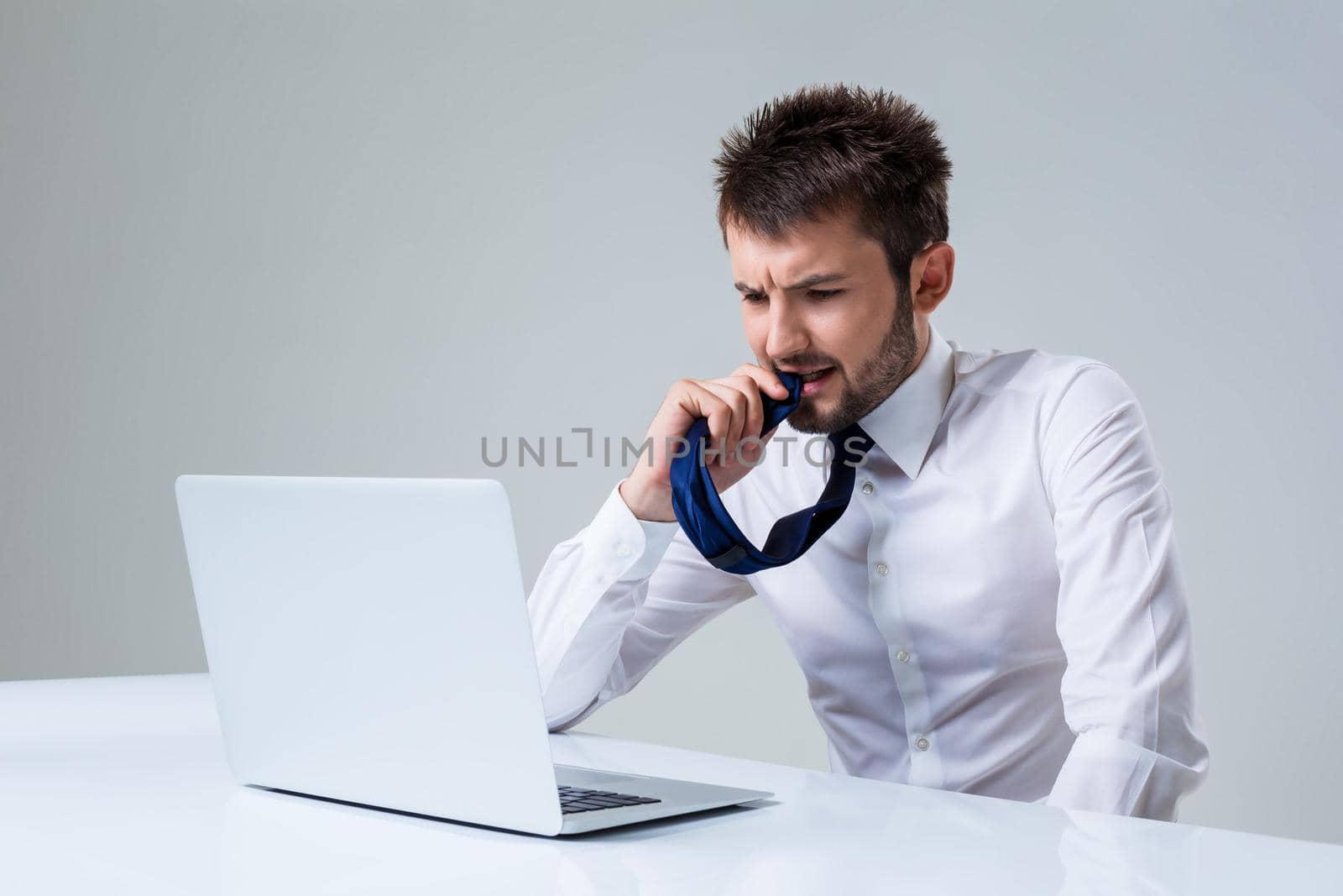 the young man is nervous and chewing on his tie using laptop computer sitting at the table. Office clothing