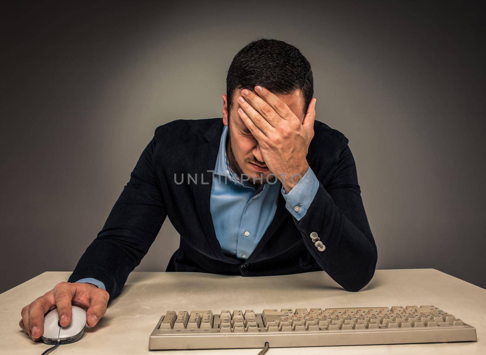 Portrait frustrated young man covering face with hands while sitting at a desk near a computer, isolated on gray background. Feeling sick and tired - Human emotion.