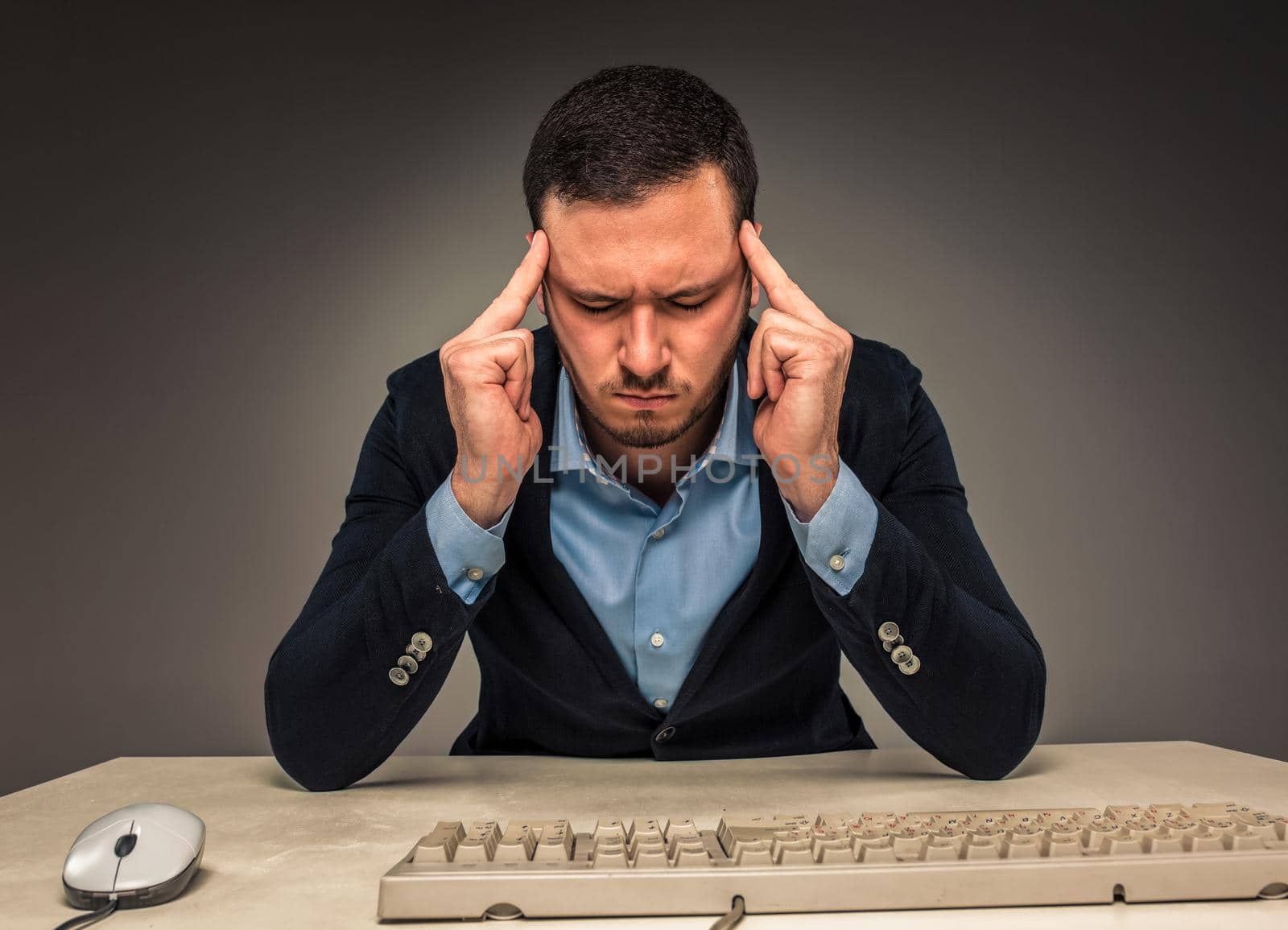 Portrait frustrated young man, hands touch the head, sitting at a desk near a computer, isolated on gray background. Feeling sick and tired - Human emotion.