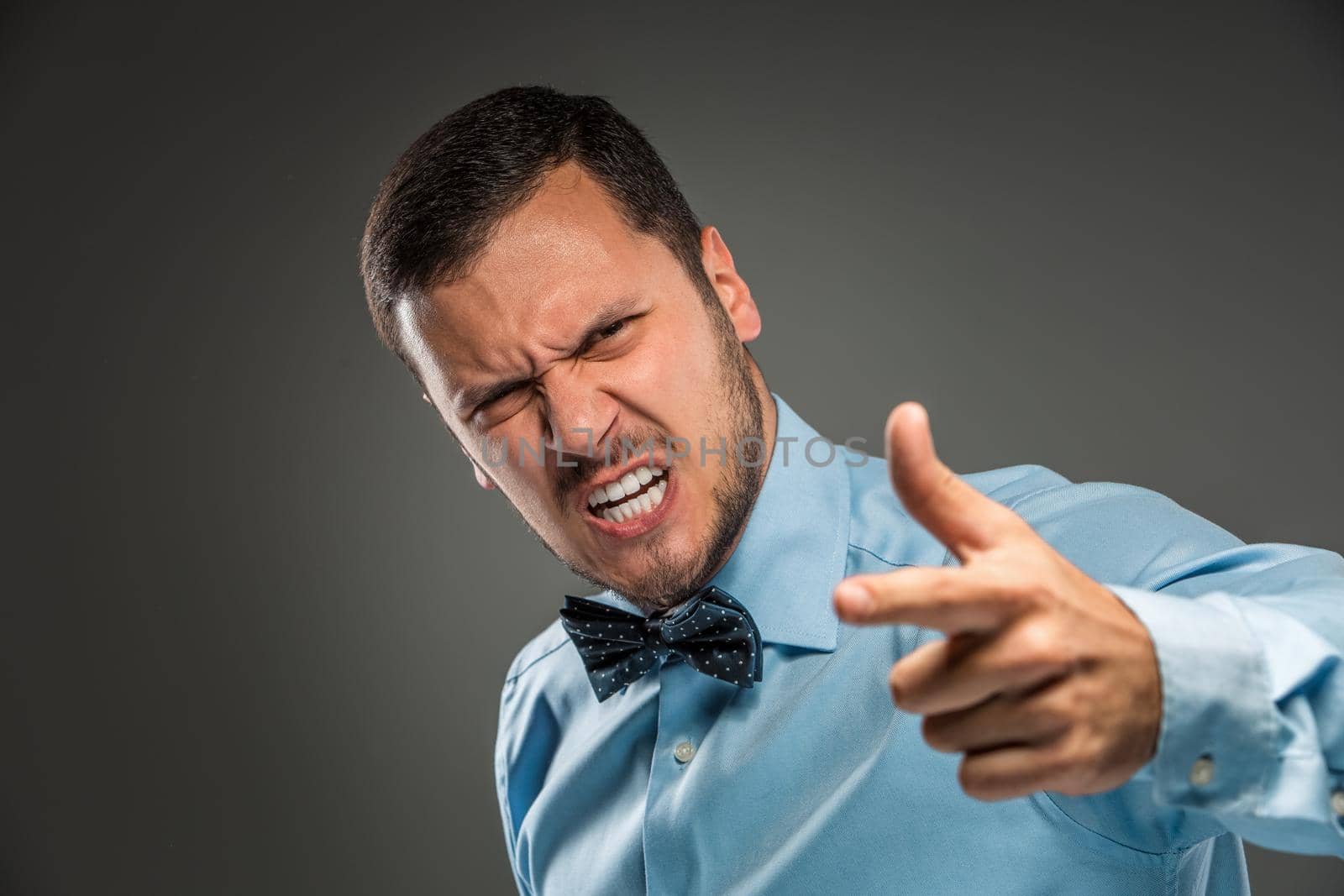 Portrait of angry upset young man in blue shirt and butterfly tie, pointing finger at camera, isolated on gray background. Negative human emotion, facial expression. Closeup