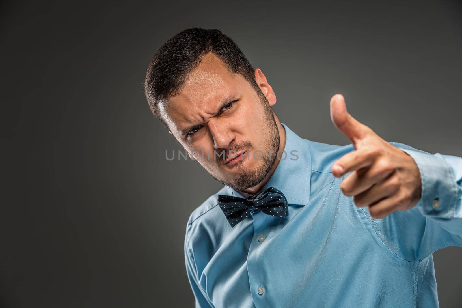 Portrait of angry upset young man in blue shirt and butterfly tie with hands up, pointing finger at camera, isolated on gray background. Negative human emotion, facial expression. Closeup