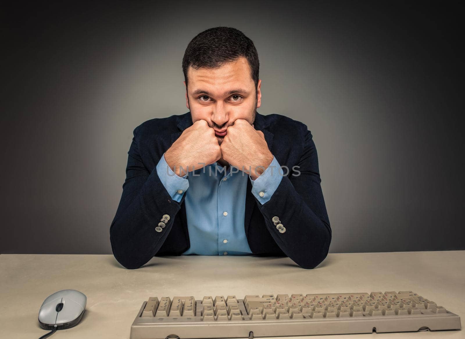 Portrait young man in blue shirt and jacket looking at the camera, hands holding his head, sitting at a desk near a computer isolated on gray studio background. Human emotion, facial expression. Closeup