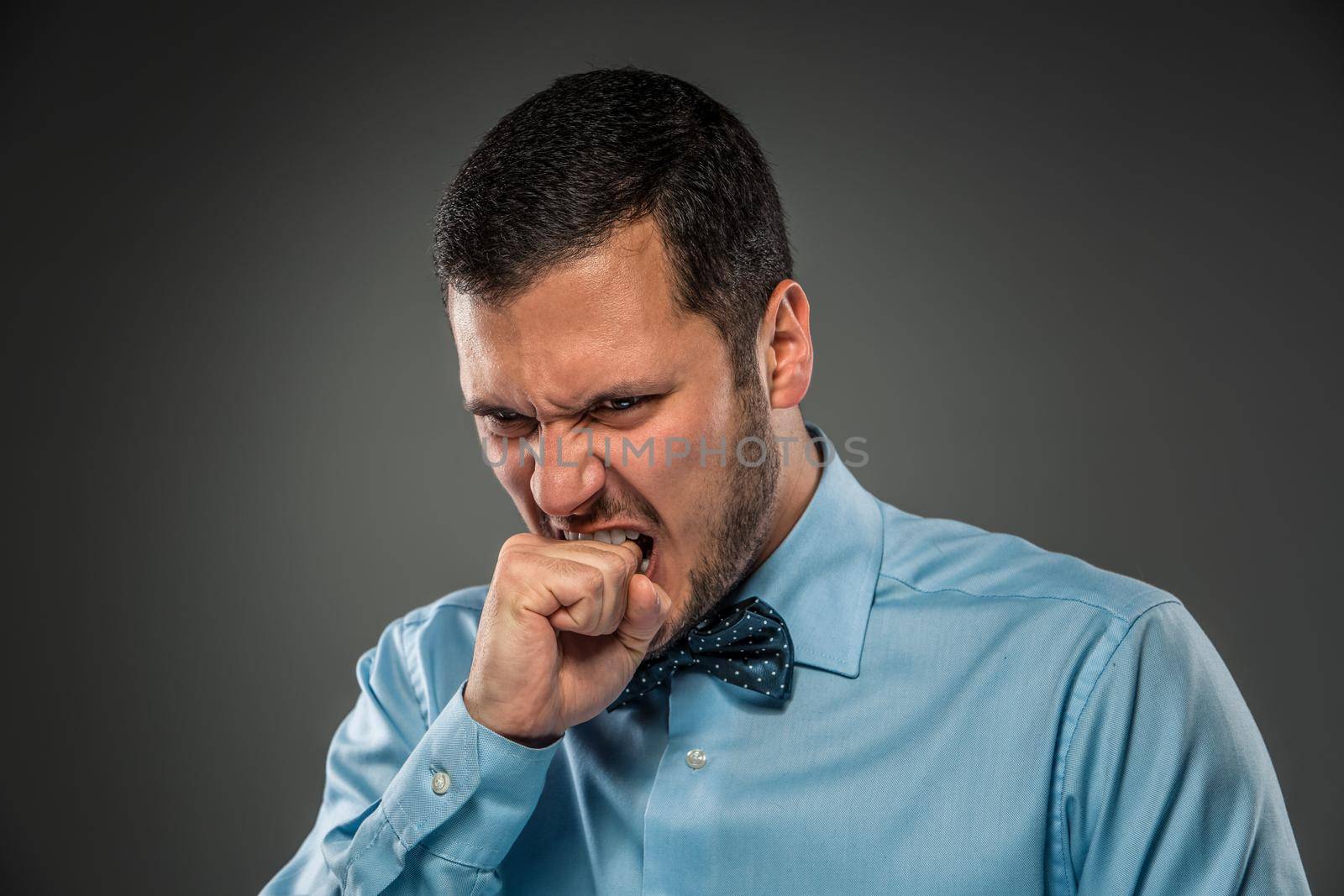 Closeup portrait of an angry guy in blue shirt and butterfly tie, biting his fist, isolated on gray background. Human emotion.