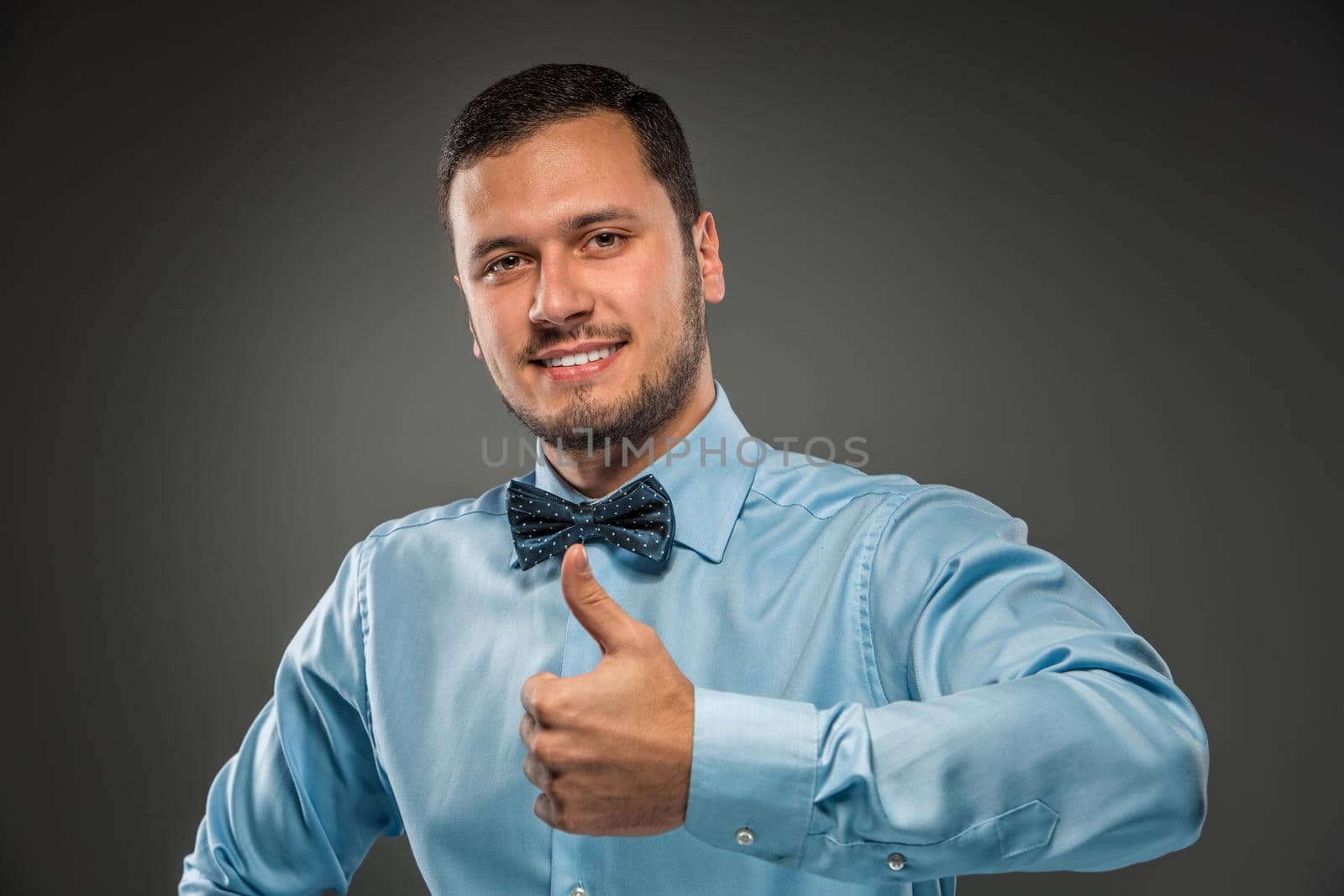 Happy smiling young man in blue shirt and butterfly tie making the ok thumbs up hand sign, gesture, isolated over gray studio background.