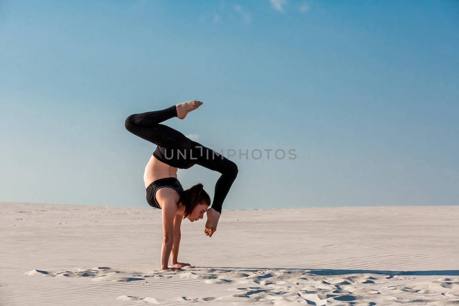 Young woman practicing inversion balancing yoga pose handstand on beach with white sand and bright blue sky