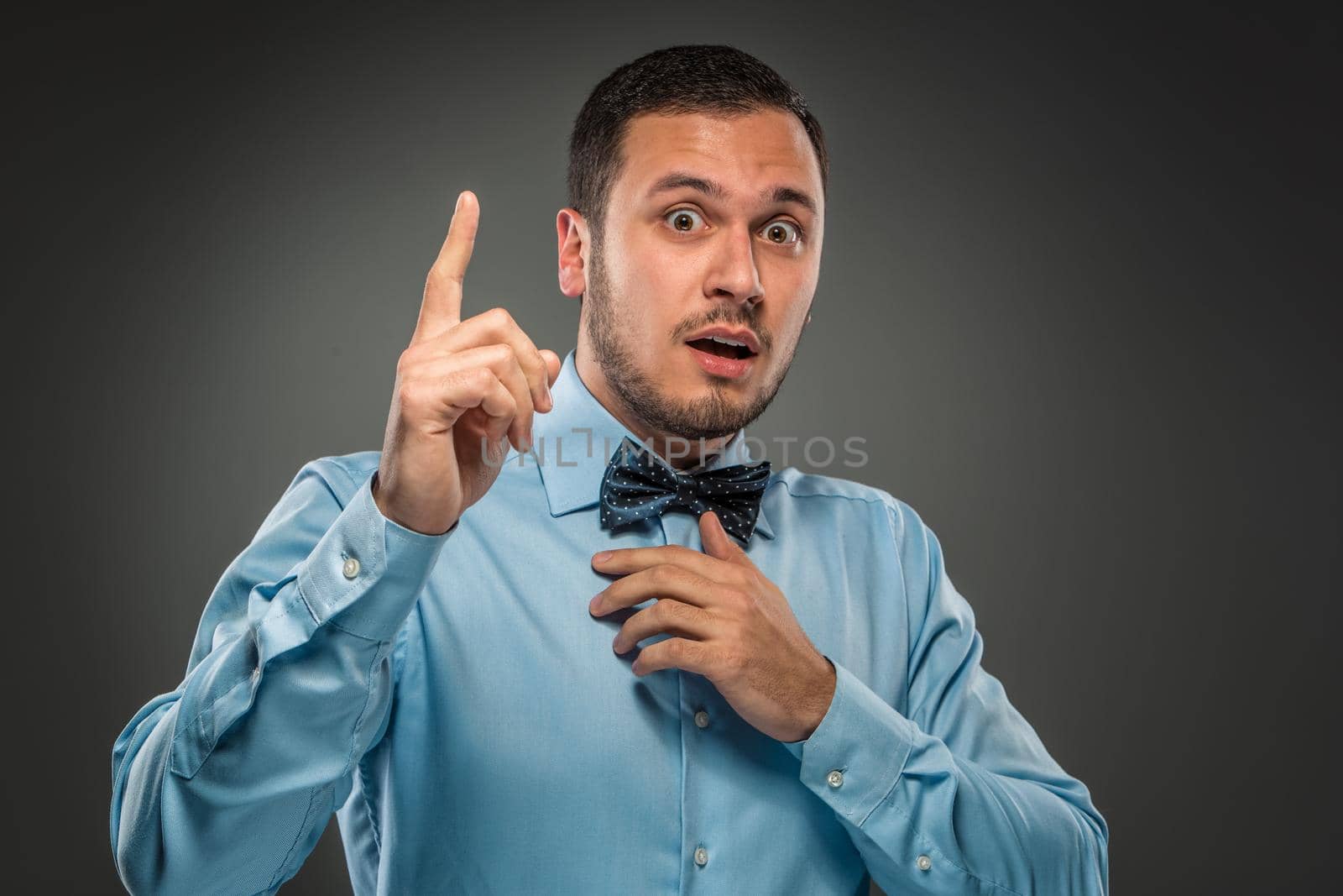 Handsome man in blue shirt and butterfly tie raised his index finger and looking at the camera isolated on gray background. Concept of the idea or warning