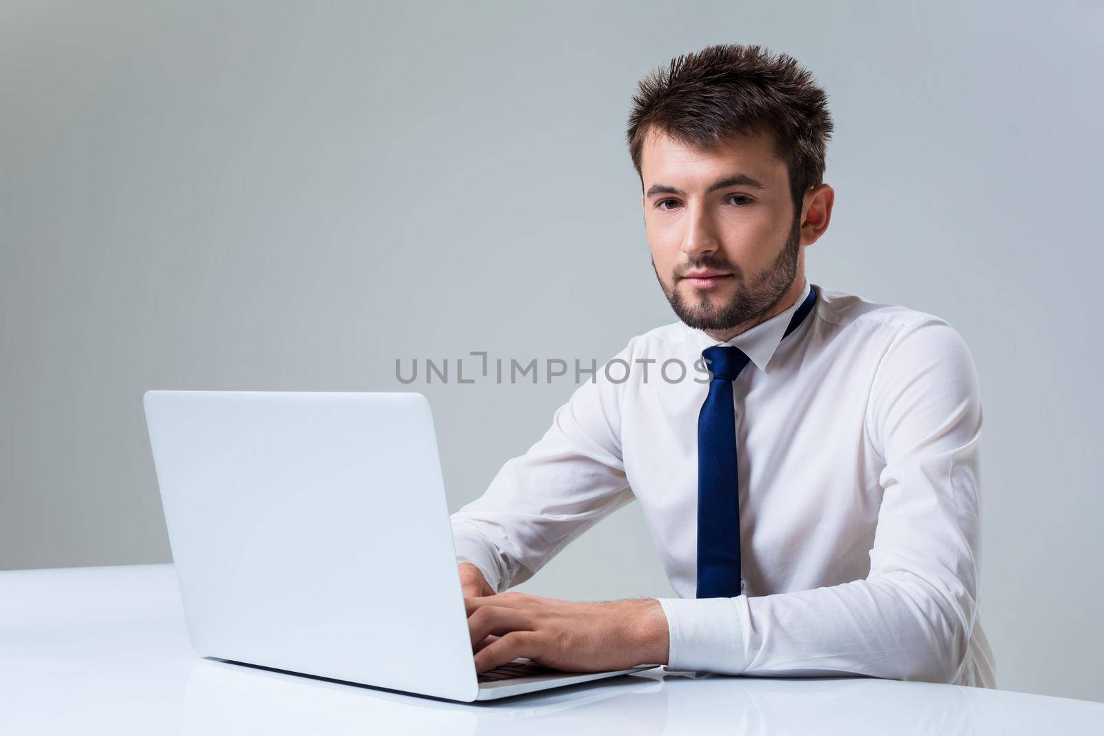 young man smiling at the camera uses a laptop computer while sitting at a table. Office clothing