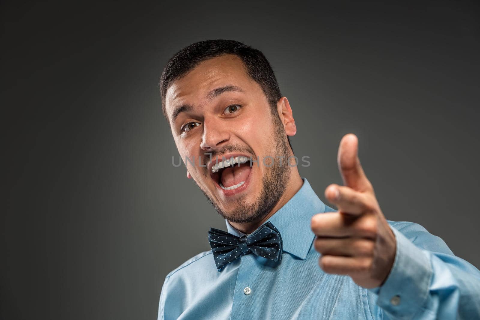 Closeup portrait of excited, energetic, happy, smiling man in blue shirt and butterfly tie is gesturing with his hand, pointing finger at camera isolated on gray background. Positive human emotion facial expression