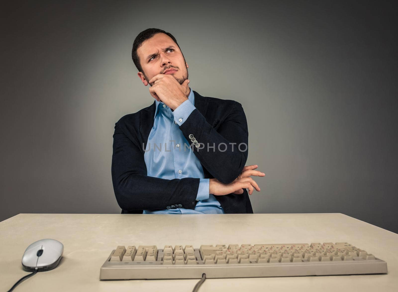 Handsome man in blue shirt and jacket looking up, sitting at a desk near a computer, isolated on gray background. Concept of the idea or warning