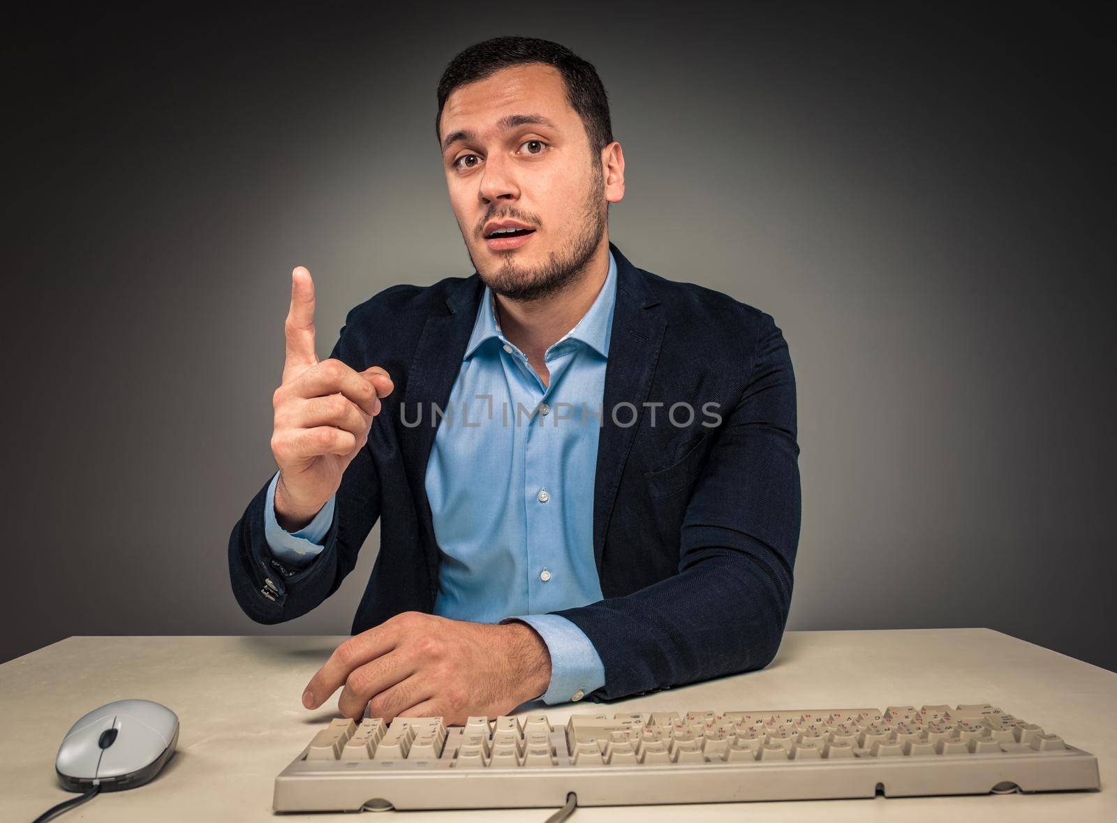 Handsome man raised his index finger and looking at the camera, sitting at a desk near a computer, isolated on gray background. Concept of the idea or warning