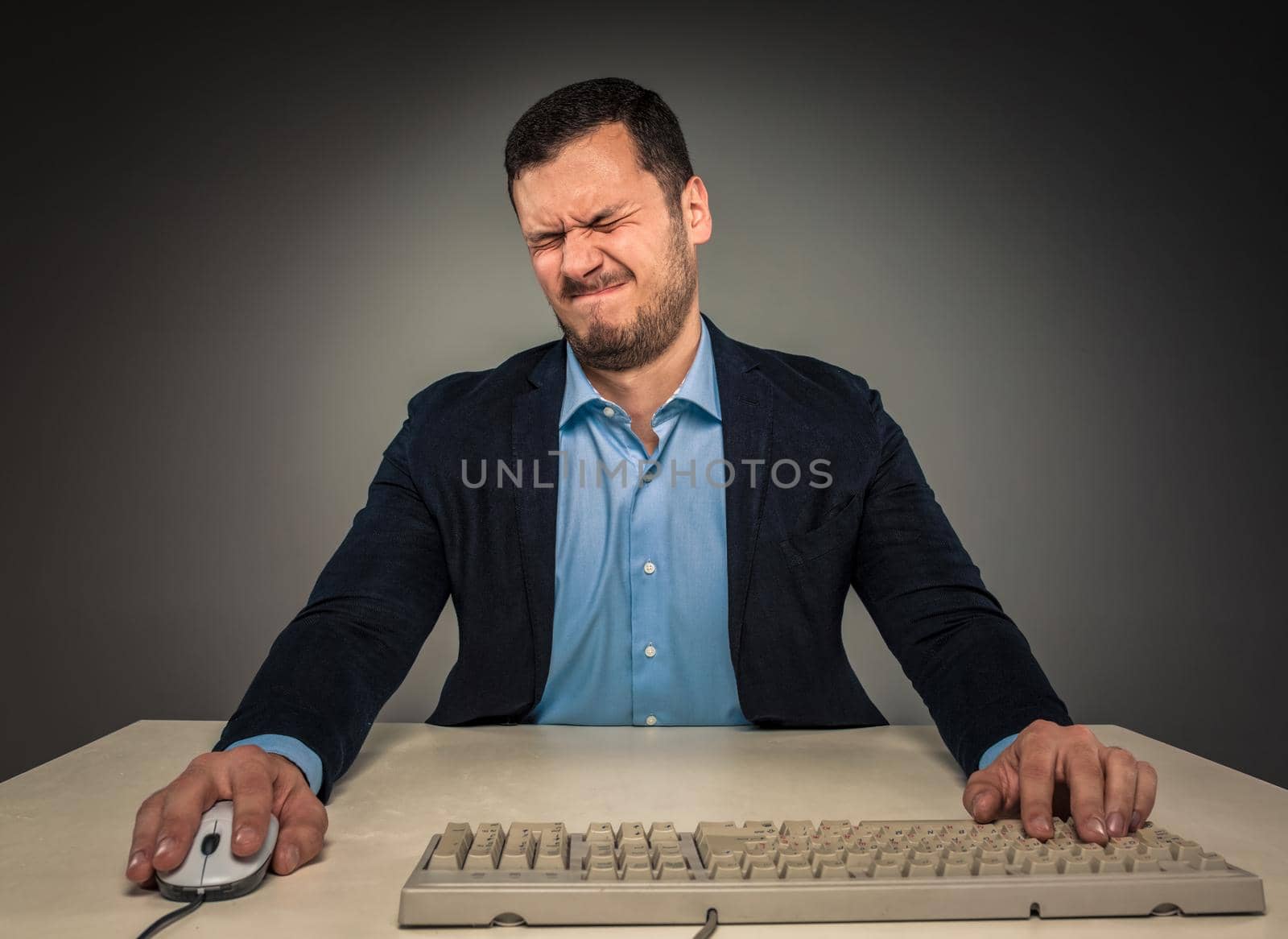 Portrait frustrated young man, with closed eyes and pursed lips, sitting at a desk near a computer, isolated on gray background. Feeling sick and tired - Human emotion.