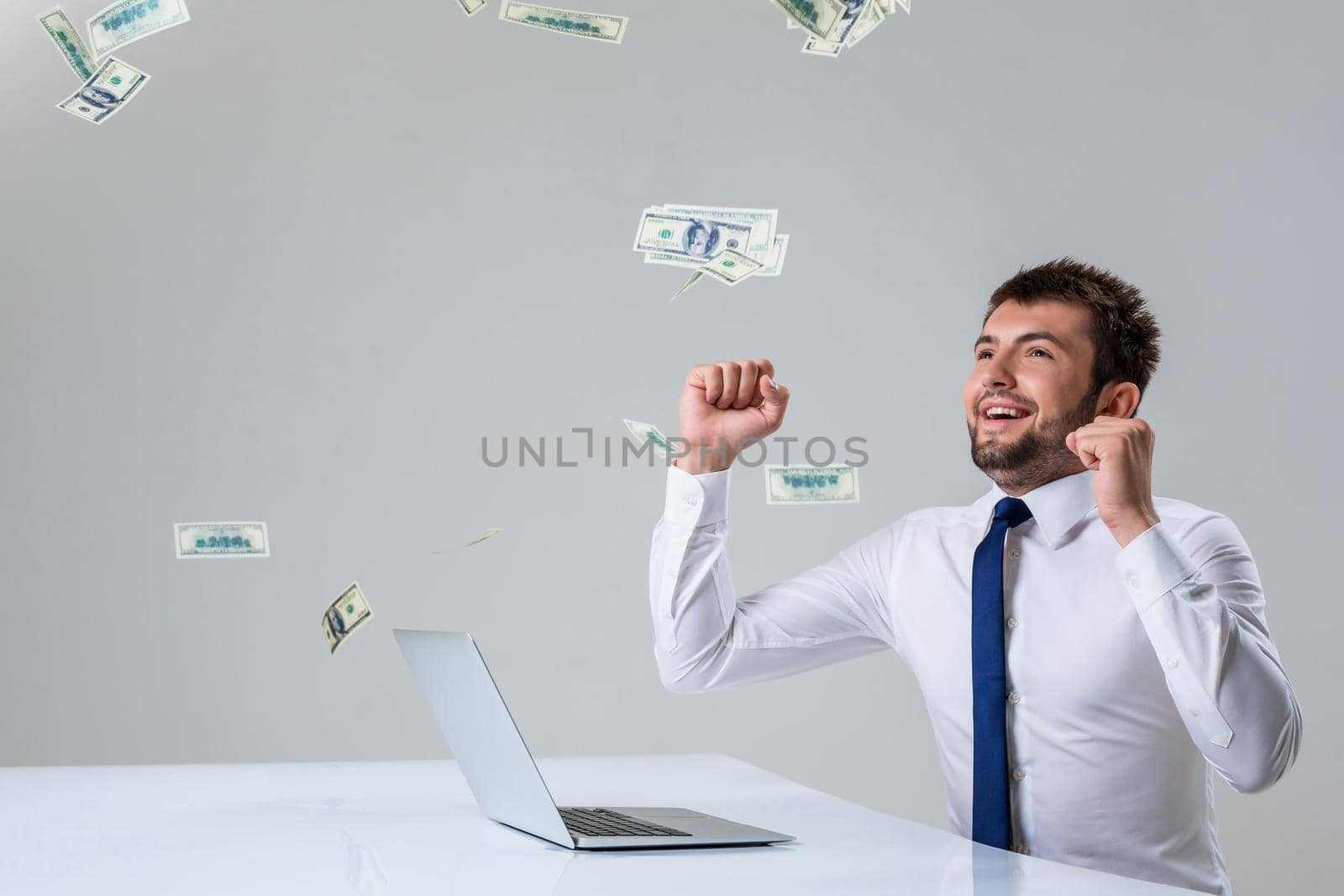 the young man behind the laptop. It uses a computer while sitting at a table. enjoys hands up. Office clothing. the top falling money, dollars