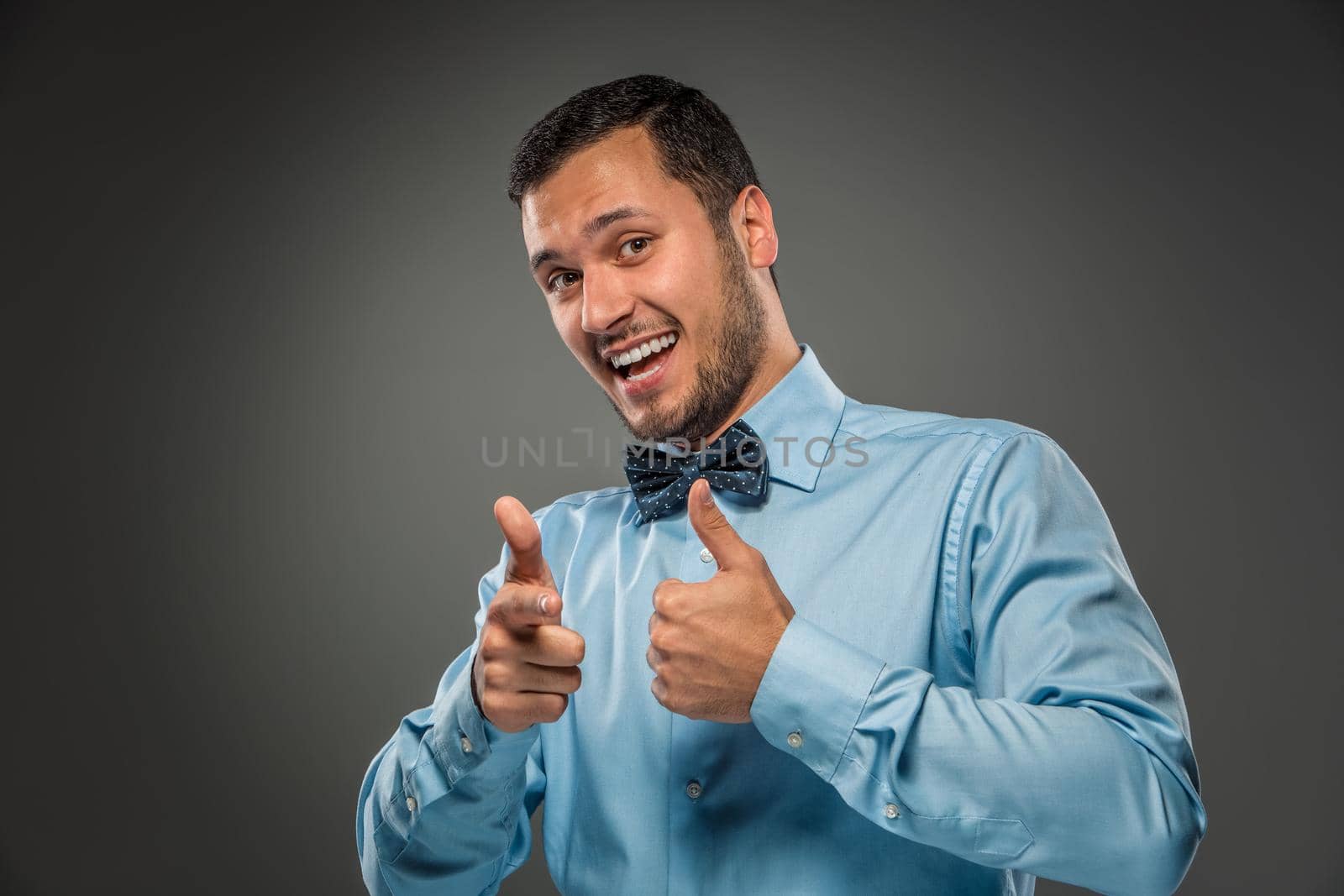 Closeup portrait of excited, energetic, happy, smiling man in blue shirt and butterfly tie is gesturing with his hand, pointing finger at camera isolated on gray background. Positive human emotion facial expression