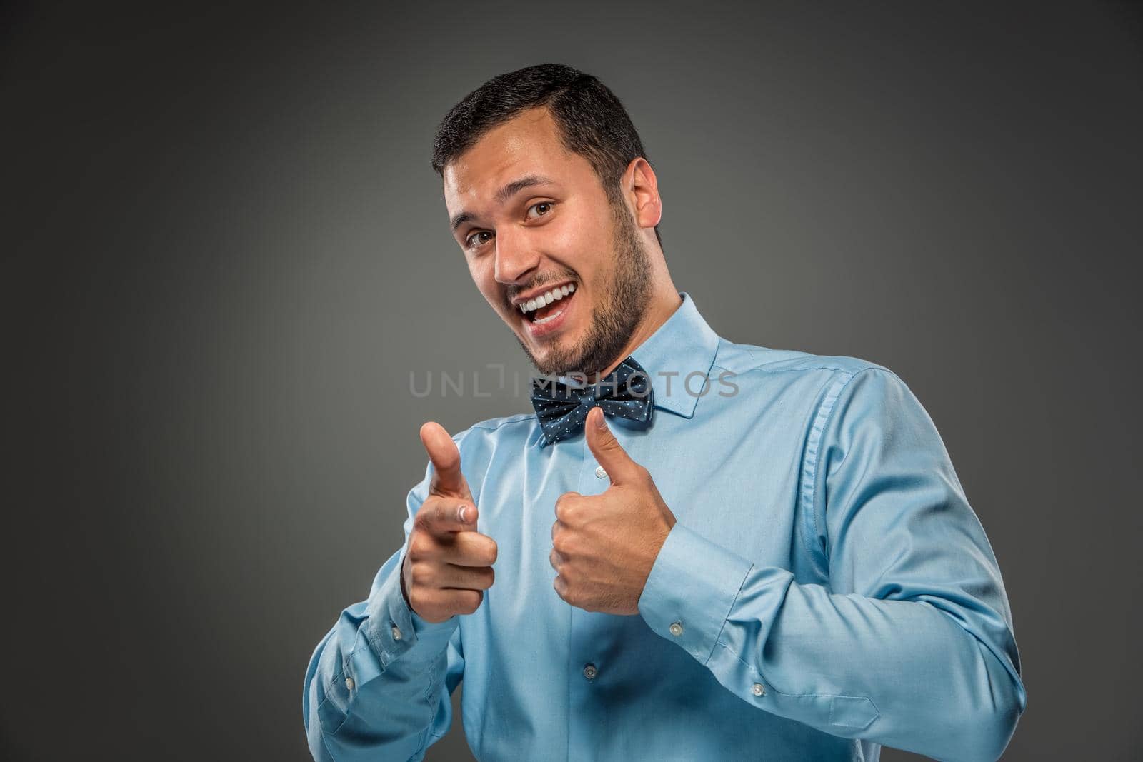 Closeup portrait of excited, energetic, happy, smiling man in blue shirt and butterfly tie is gesturing with his hand, pointing finger at camera isolated on gray background. Positive human emotion facial expression