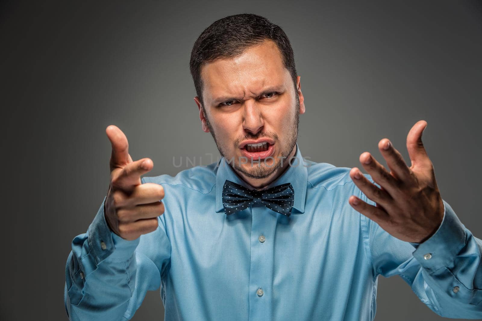 Portrait of angry upset young man in blue shirt and butterfly tie with hands up, pointing finger at camera, isolated on gray background. Negative human emotion, facial expression. Closeup