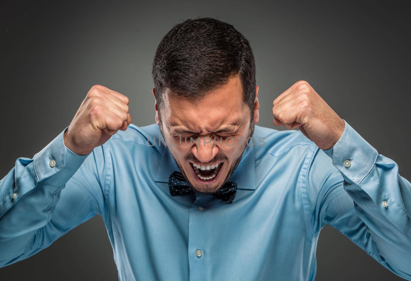 Portrait of angry upset young man in blue shirt and butterfly tie with fists up yelling isolated on gray studio background. Negative human emotion, facial expression. Closeup