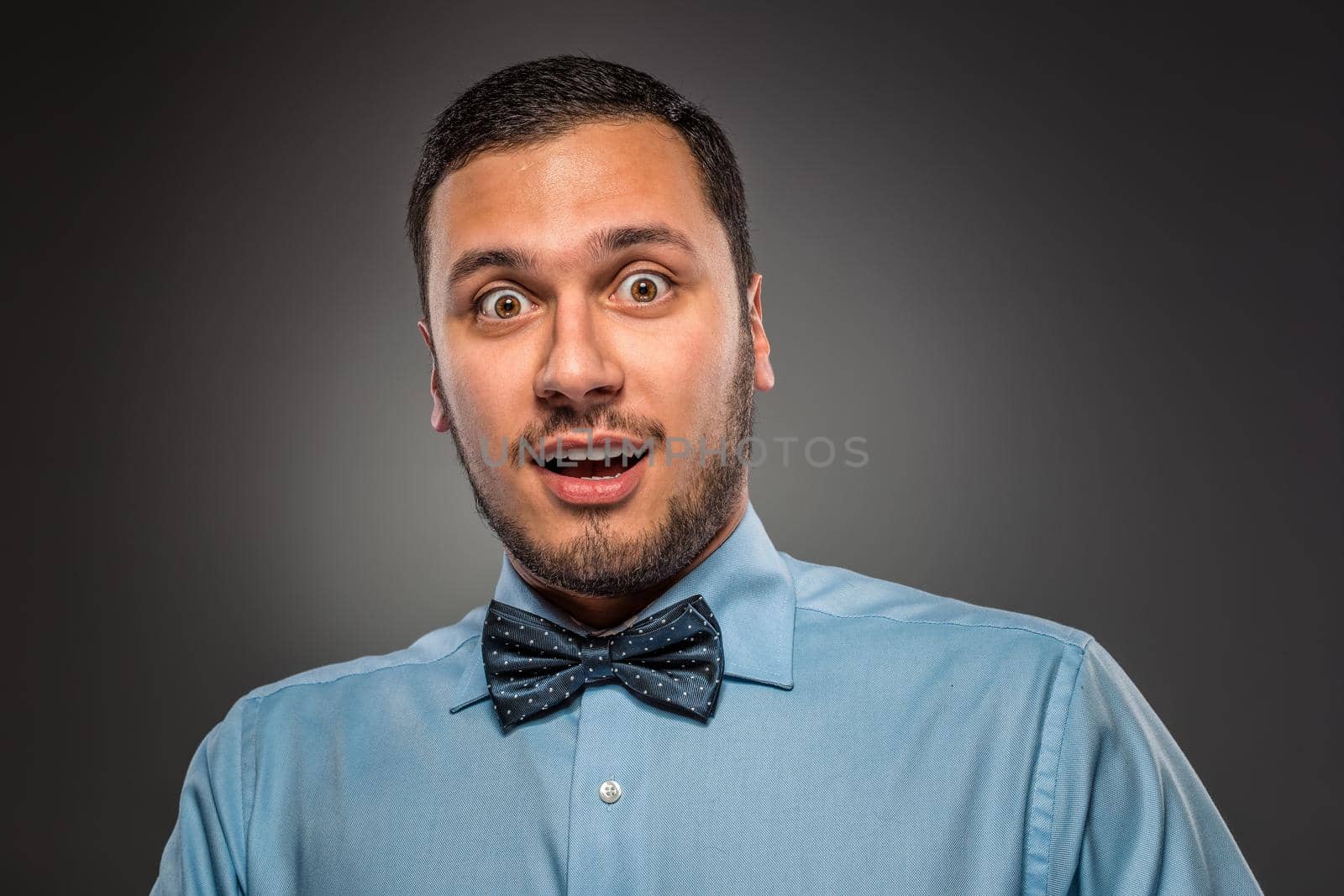 Portrait young man in blue shirt and butterfly tie looking at the camera with amazement, isolated on gray studio background. Human emotion, facial expression. Closeup