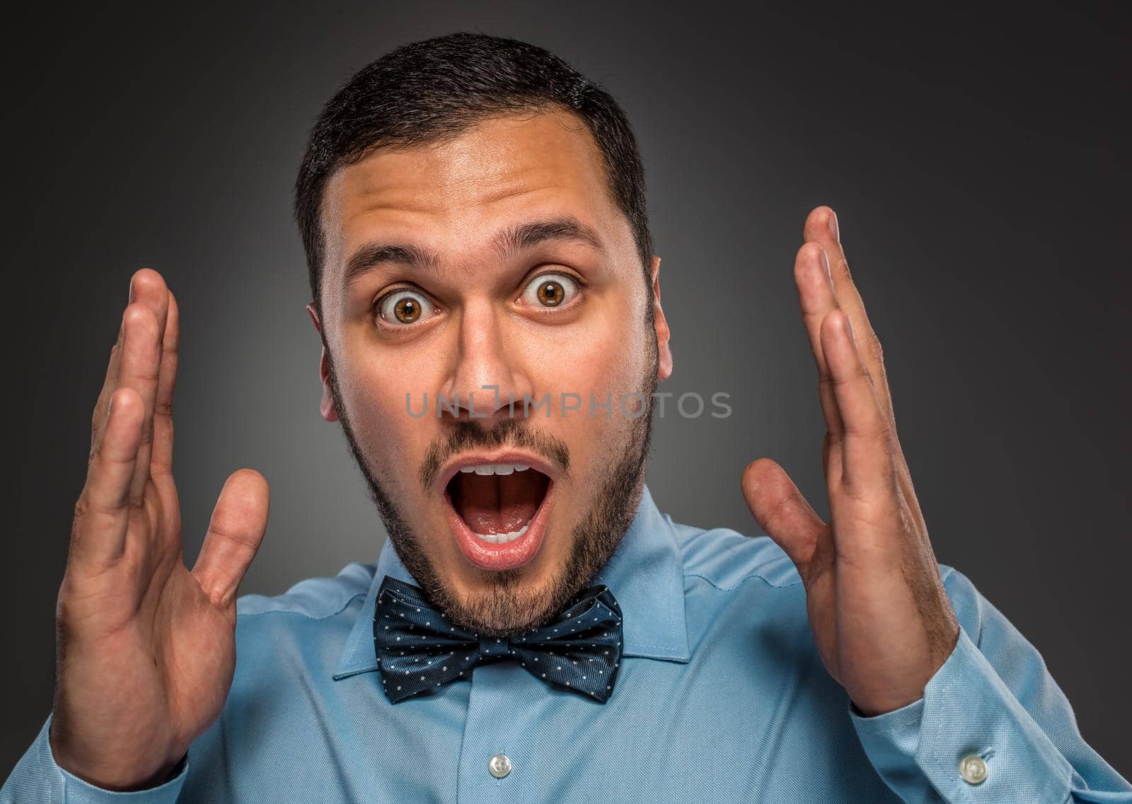 Portrait young man in blue shirt and butterfly tie looking at the camera with amazement, isolated on gray studio background. Human emotion, facial expression. Closeup