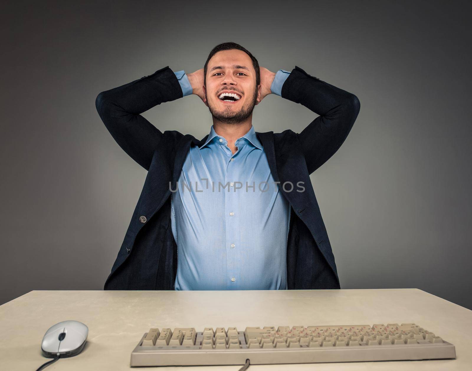 Closeup portrait of excited, energetic, happy, man in blue shirt and butterfly tie winning, his hands kept his head sitting at a desk near a computer isolated on gray background. Positive human emotion facial expression