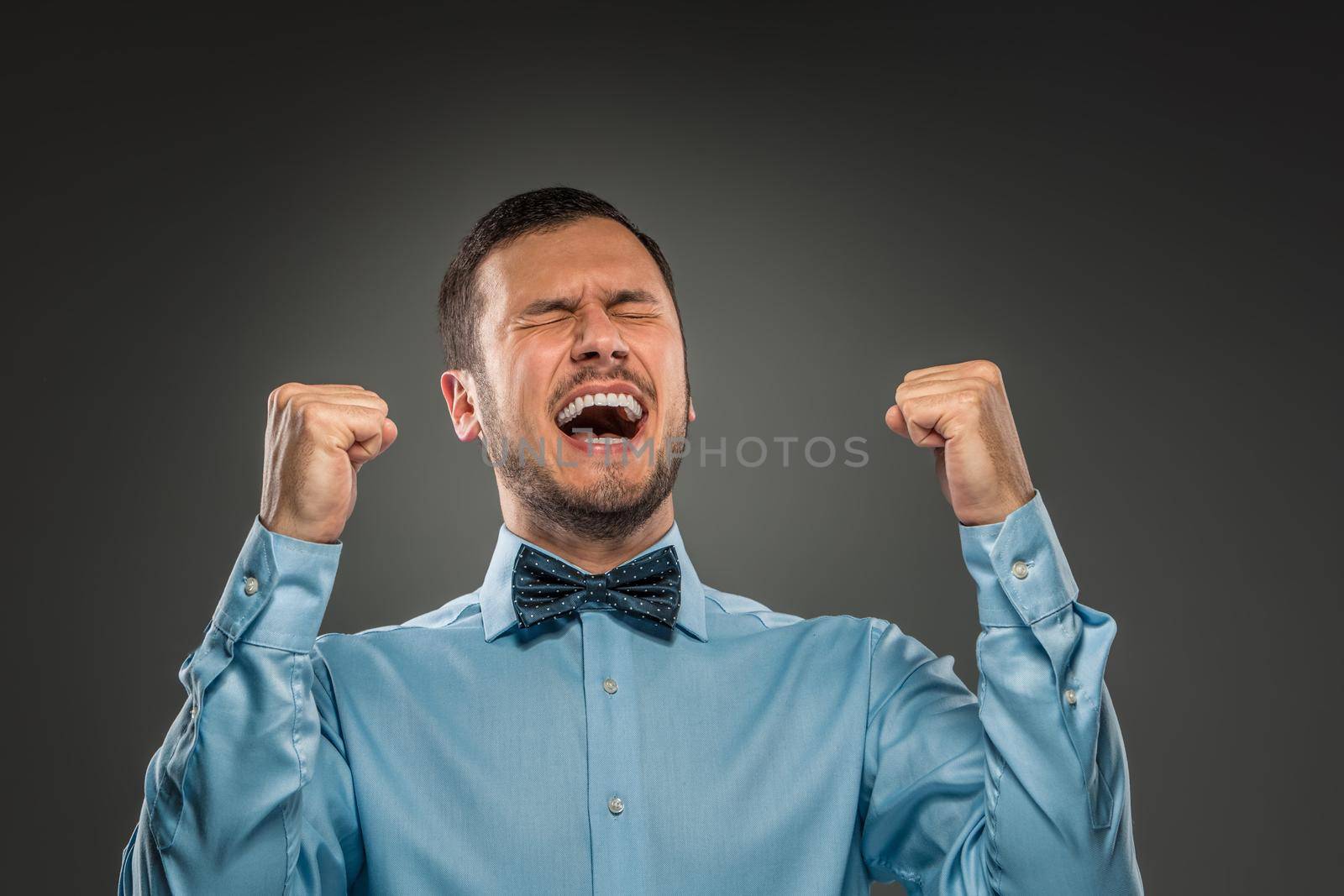 Closeup portrait of excited, energetic, happy, man in blue shirt and butterfly tie winning, arms up fists pumped celebrating success isolated grey studio background. Positive human emotion facial expression