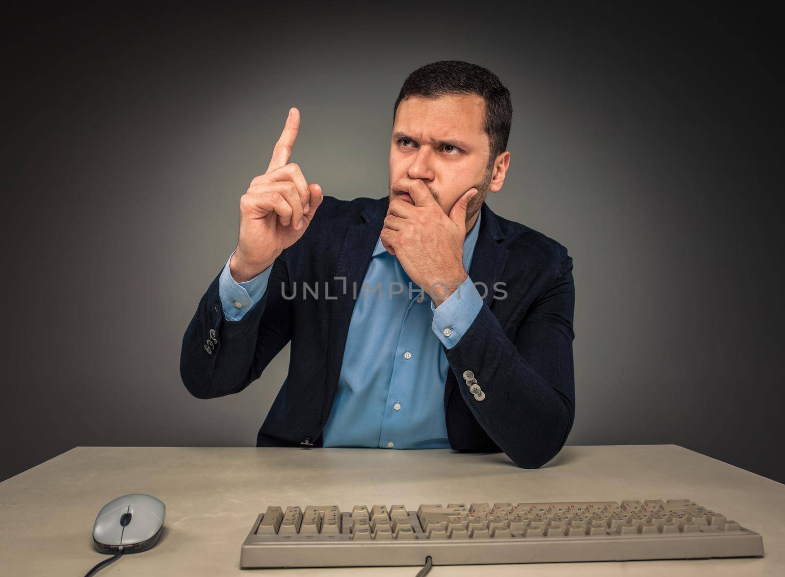 Handsome man raised his index finger and looking at the camera, sitting at a desk near a computer, isolated on gray background. Concept of the idea or warning