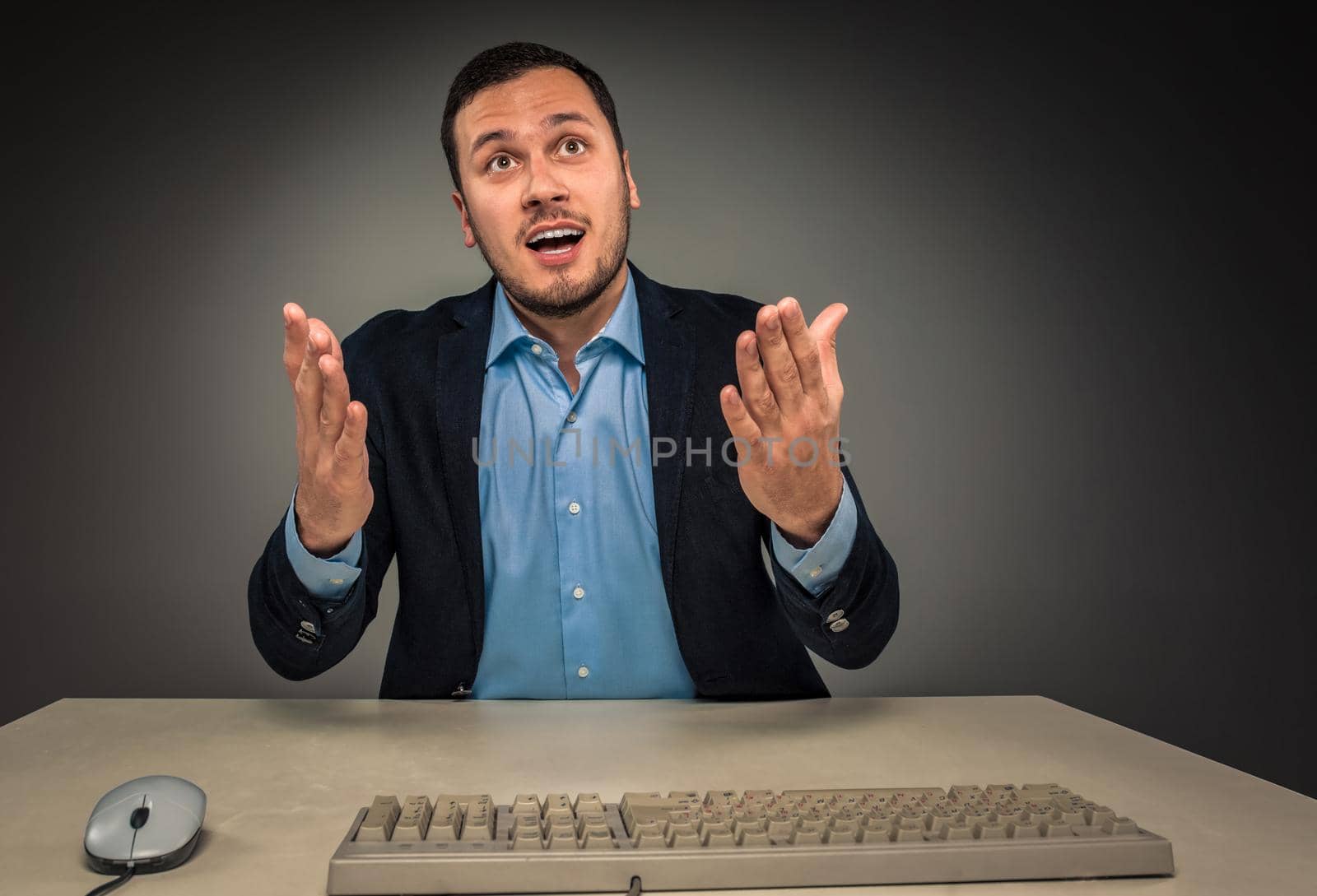 Portrait young man in blue shirt and jacket looking at the camera, hands raised, sitting at a desk near a computer isolated on gray studio background. Human emotion, facial expression. Closeup