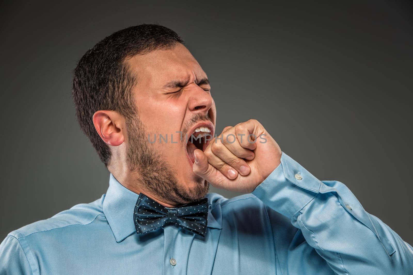 Young man in blue shirt and butterfly tie, yawns, covering her mouth isolated on gray background. Closeup
