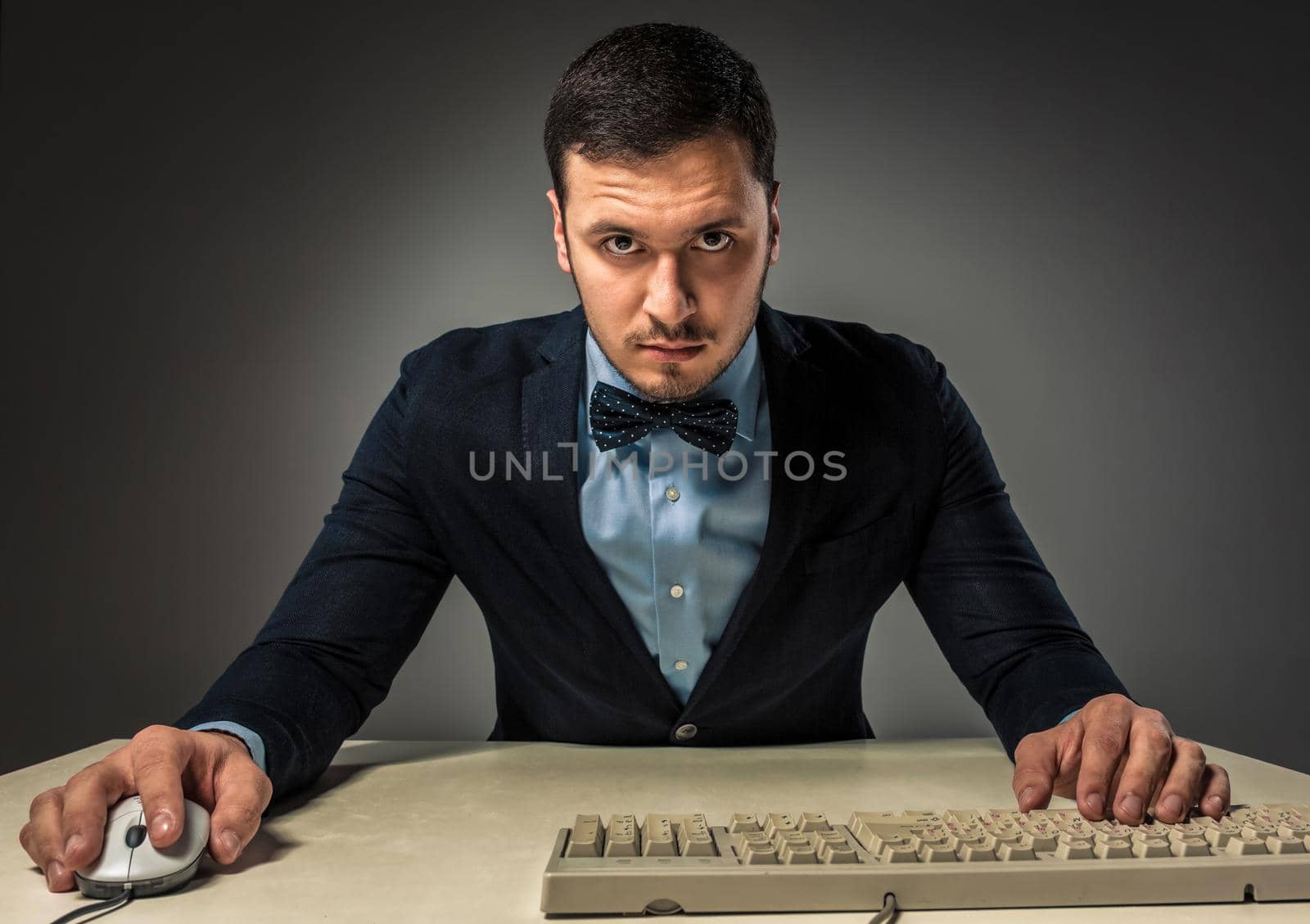 Portrait young man in blue shirt and jacket looking at the camera, sitting at a desk near a computer isolated on gray studio background. Human emotion, facial expression. Closeup