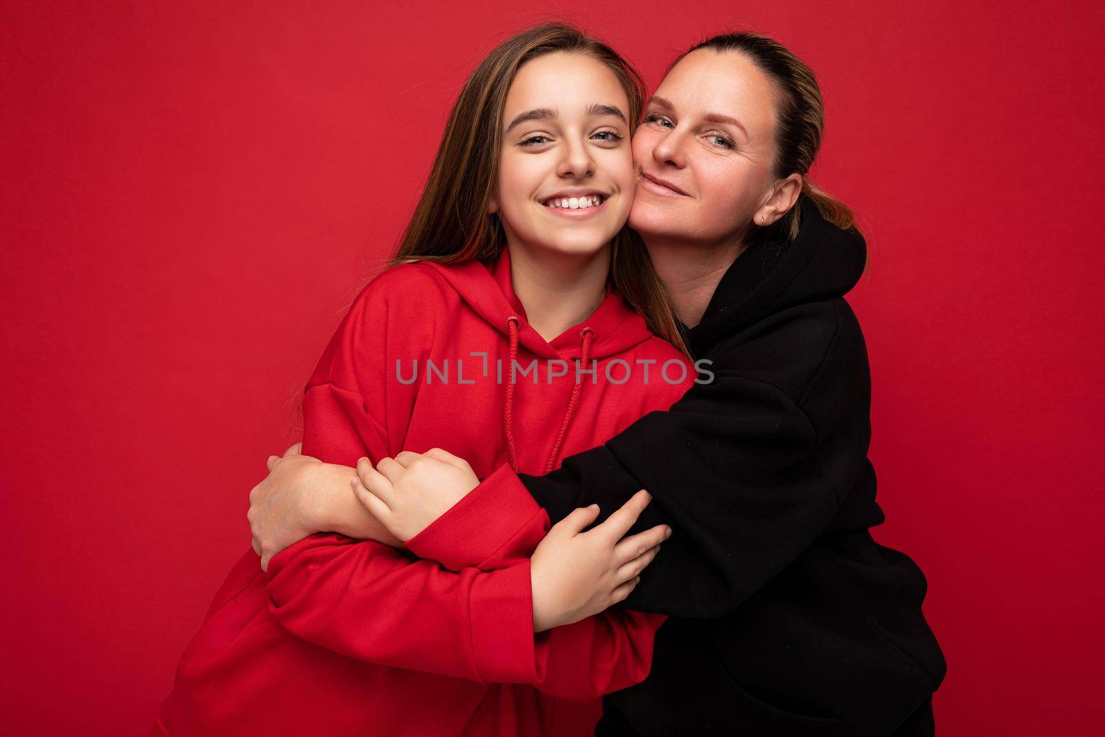 Photo of beautiful happy smiling young daughter wearing stylish red hoodie and adult mother wearing trendy black hoodie isolated over red background looking at camera and having fun by TRMK