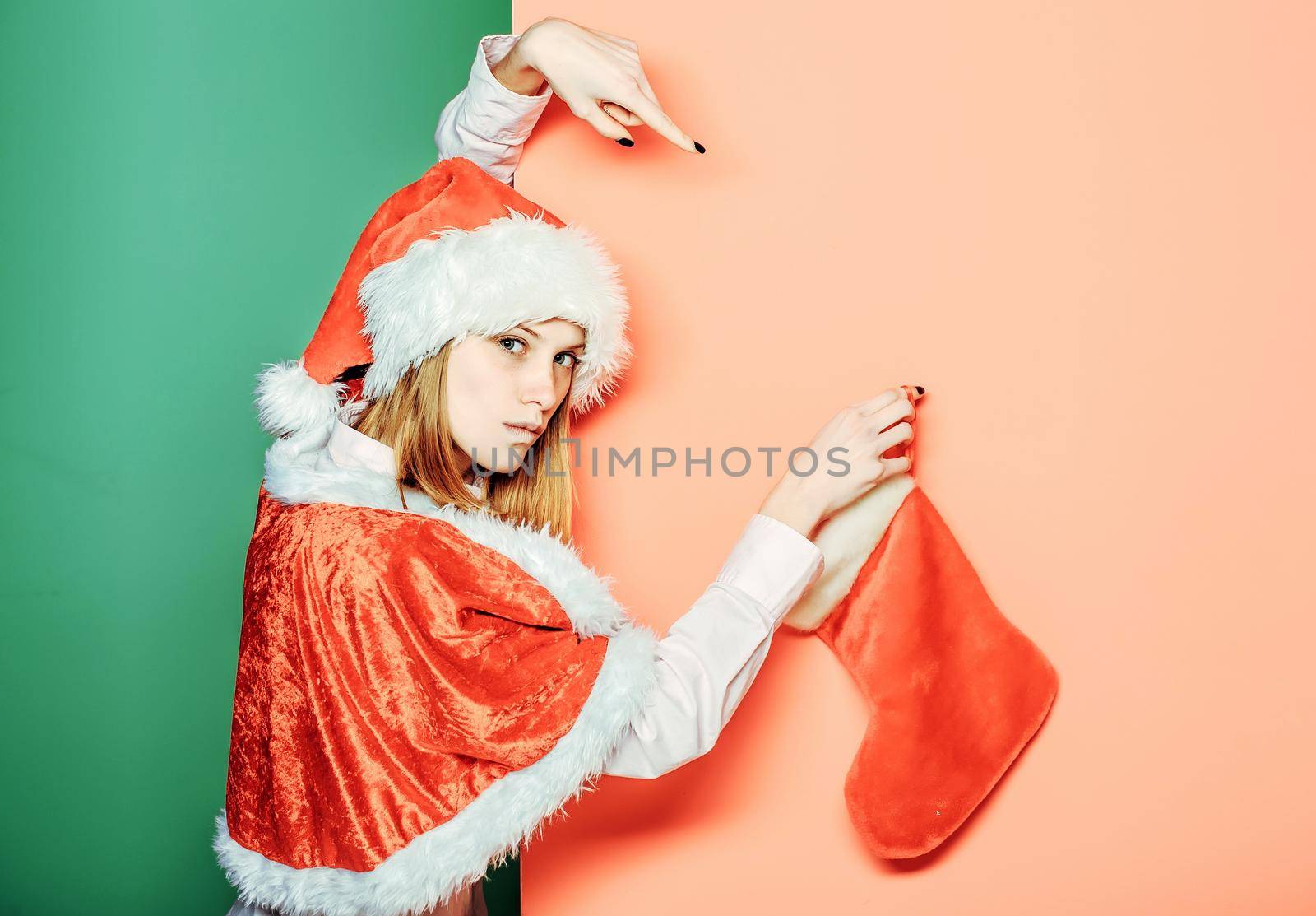 Young woman or cute serious girl with pretty face in christmas santa holds Christmas stocking on orange green isoalted background. by Tverdokhlib