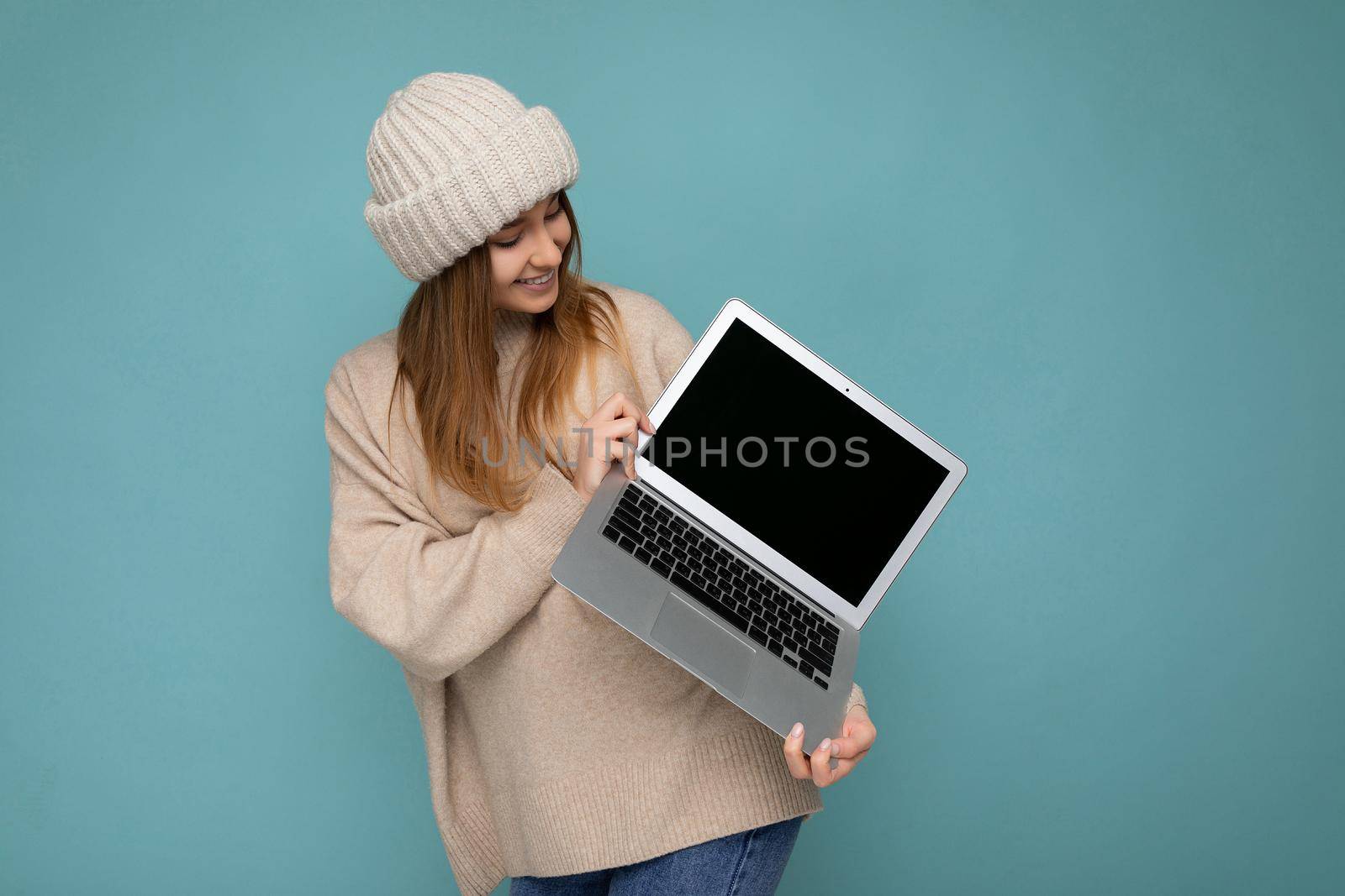 photo shot of beautiful charming fascinating pretty smiling happy young dark blond woman in winter warm knitted beige hat holding computer laptop looking at netbook empty free copy space monitor and keyboard wearing beige winter sweater isolated over light blue wall background in studio. Mock up, copy space, empty space for advertisement.