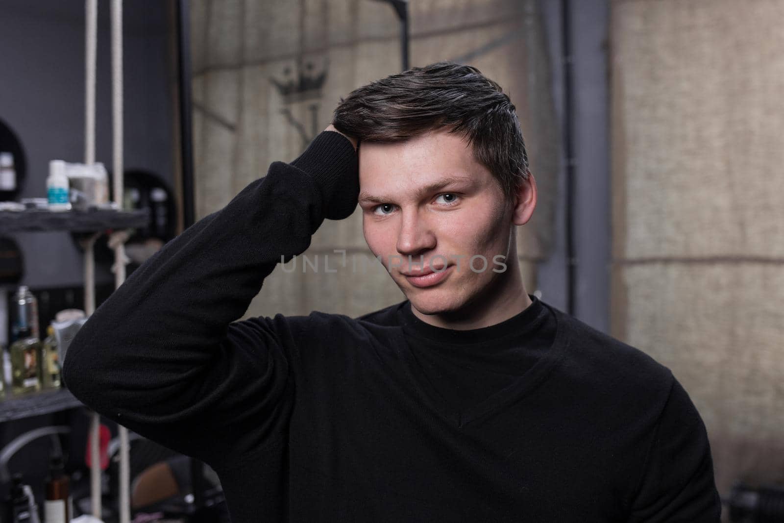 A young attractive man of Caucasian appearance in a black sweatshirt corrects his dark hair with his hand in a hairdresser's shop.