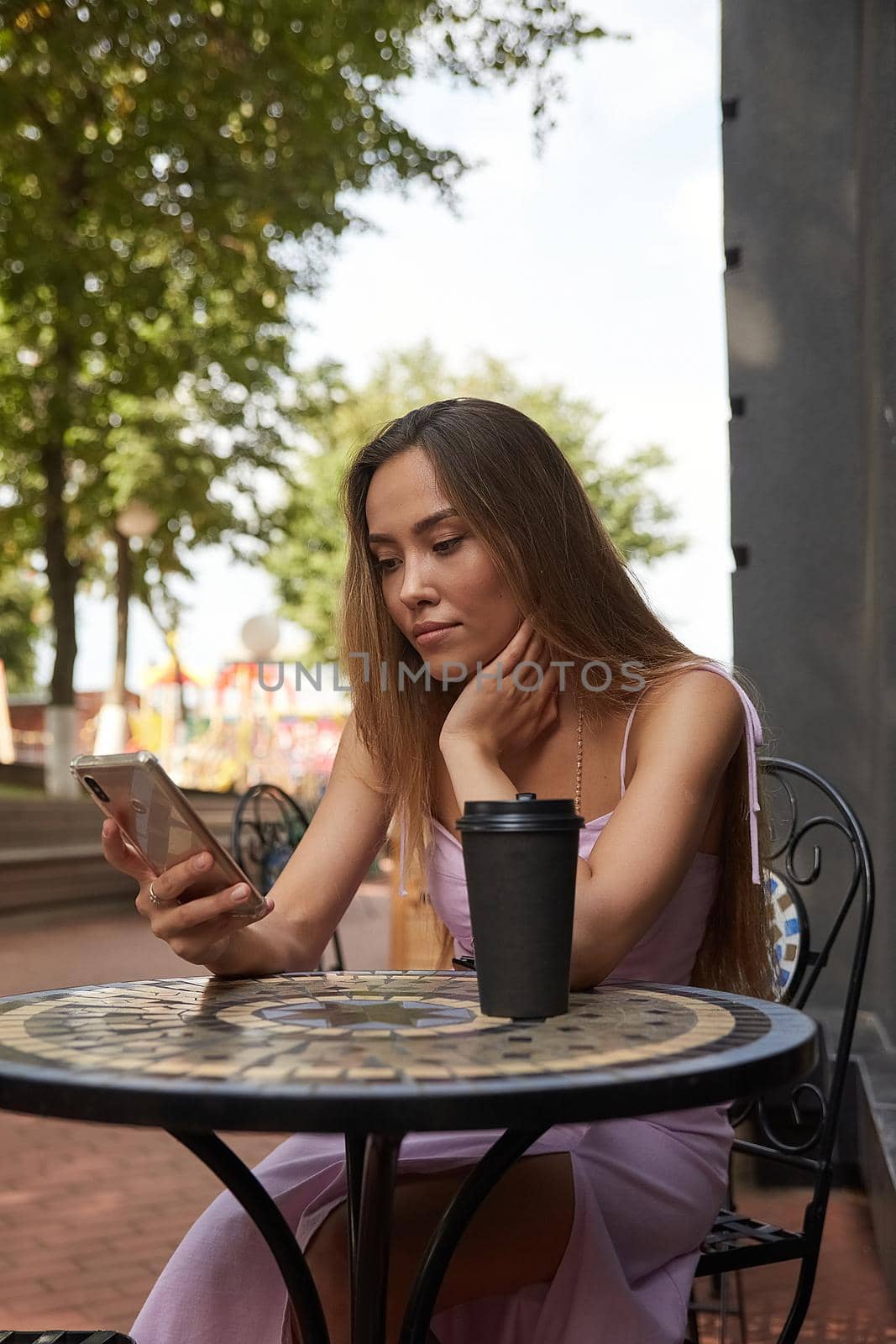 young well dressed woman sitting at table with drink, using her smartphone. lady having cup of coffee outside of cafe, surfing internet on cell phone, chatting online. modern communication technology
