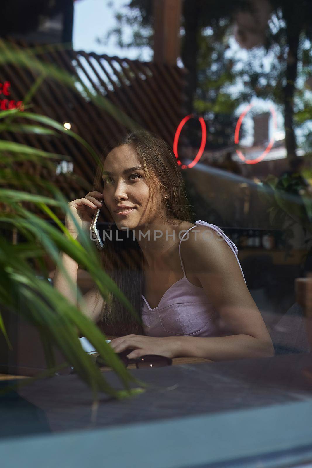 smiling pretty lady calling by phone shot through window glass outside of cafe by artemzatsepilin
