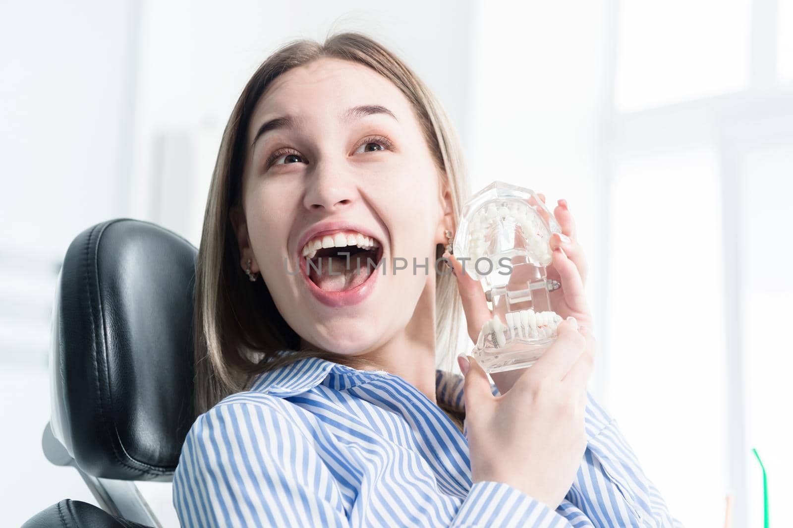 Portrait of a happy attractive girl in a dental chair. Laughing girl at the dentist's appointment with a jaw model in her hands.