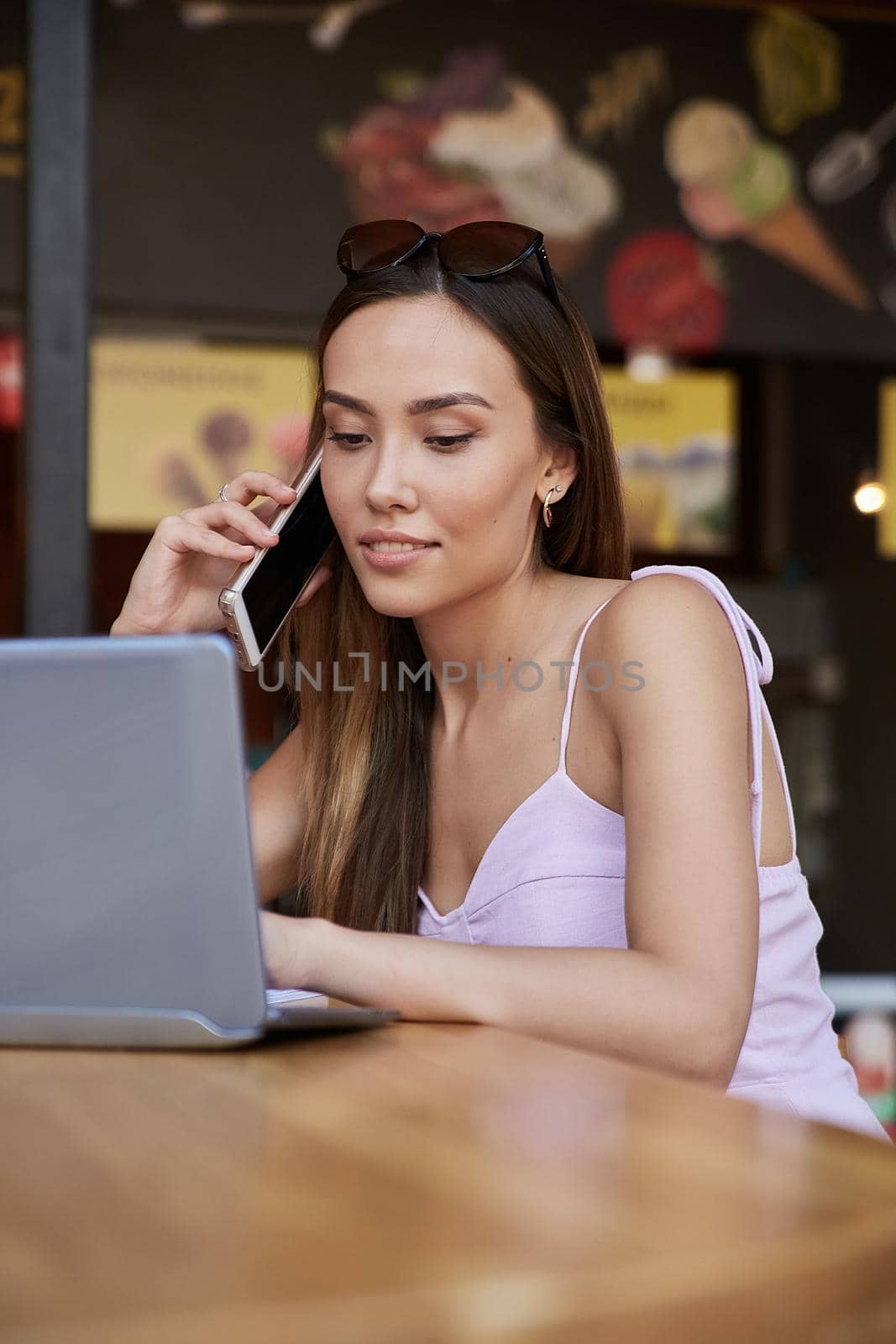young asian pretty woman in pink dress sitting at diner, using laptop, calling by smartphone. attractive female in light pink dress working at cafe. lifestyle portrait, modern communication