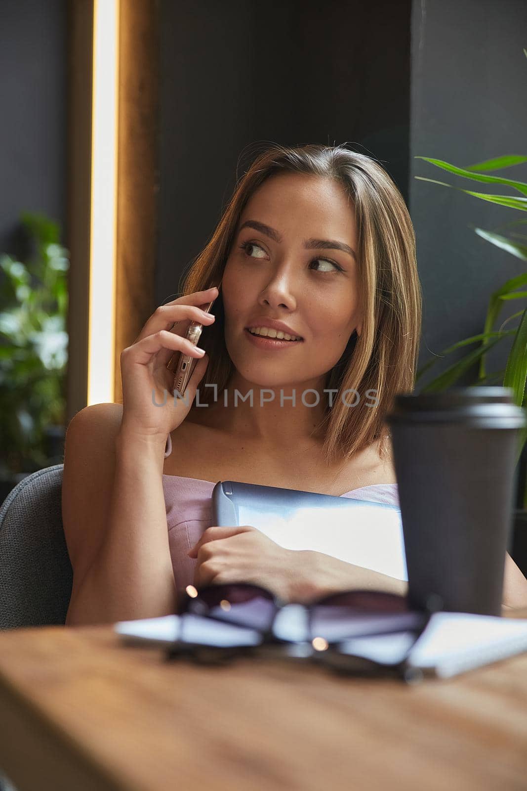 young asian pretty woman sitting at table of coffee shop, calling by smartphone. beautiful lady talking by cell phone at cafe, holding laptop. modern communication technology, distant work, remote job