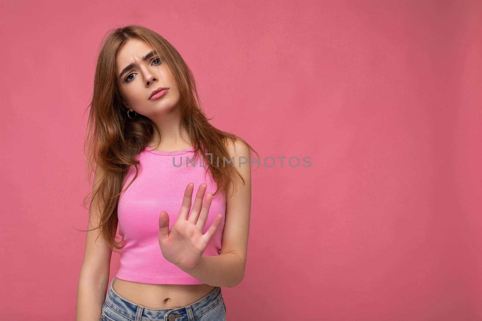 Serious upset young attractive dark blonde woman with sincere emotions isolated on background wall with copy space wearing stylish pink top and doing stop gesture. Negative concept.