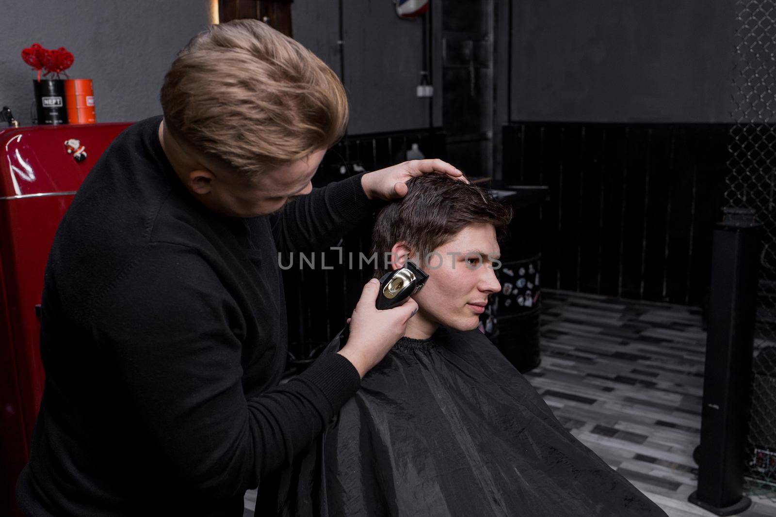 The hand of a barber or hairdresser cuts the dark long hair of the guy's client on his temple. Hairdressing.