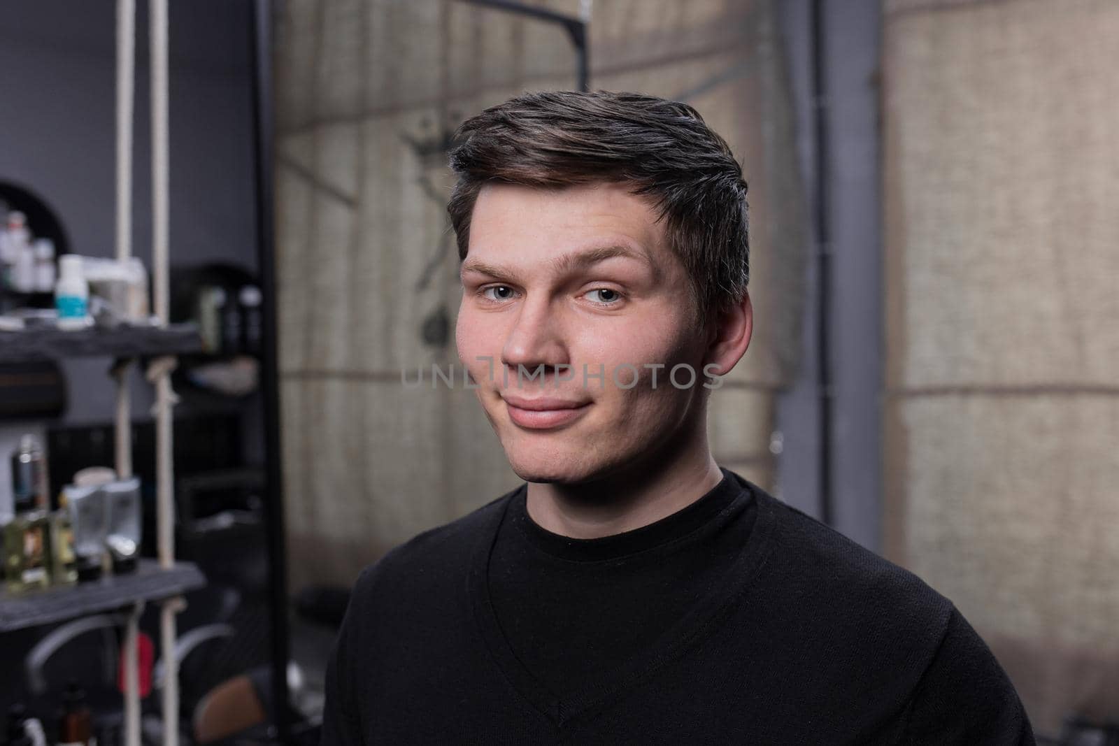 Portrait of a young positive guy of European appearance with dark hair in a black sweatshirt against the background of a hairdresser.