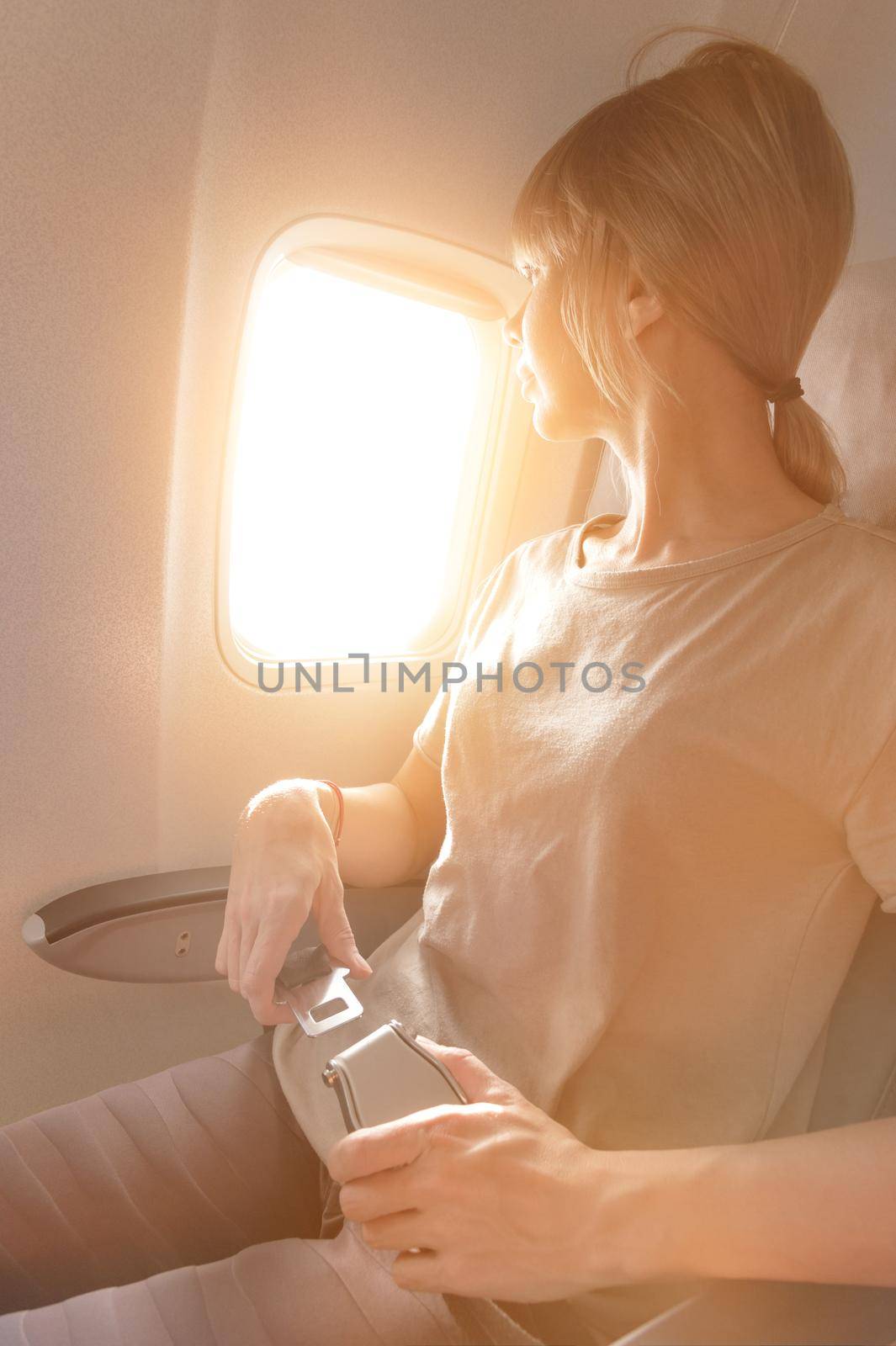 Attractive caucasian blonde girl sitting in an airplane chair looking out the window and fastens her seat belt. Safe Air Transport Concept by yanik88