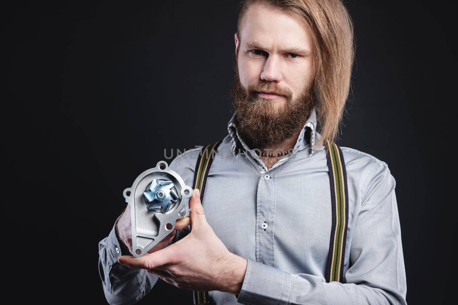 Studio portrait of a bearded stylish long-haired man in a shirt with suspenders. Holding car parts internal combustion engine water cooling pump