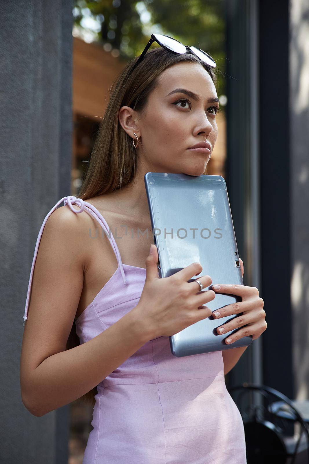 beautiful young asian woman in pink dress with dark long hair, standing outdoors holding tablet. attractive pensive female having rest outside of cafe. modern communication, social media, summer chill