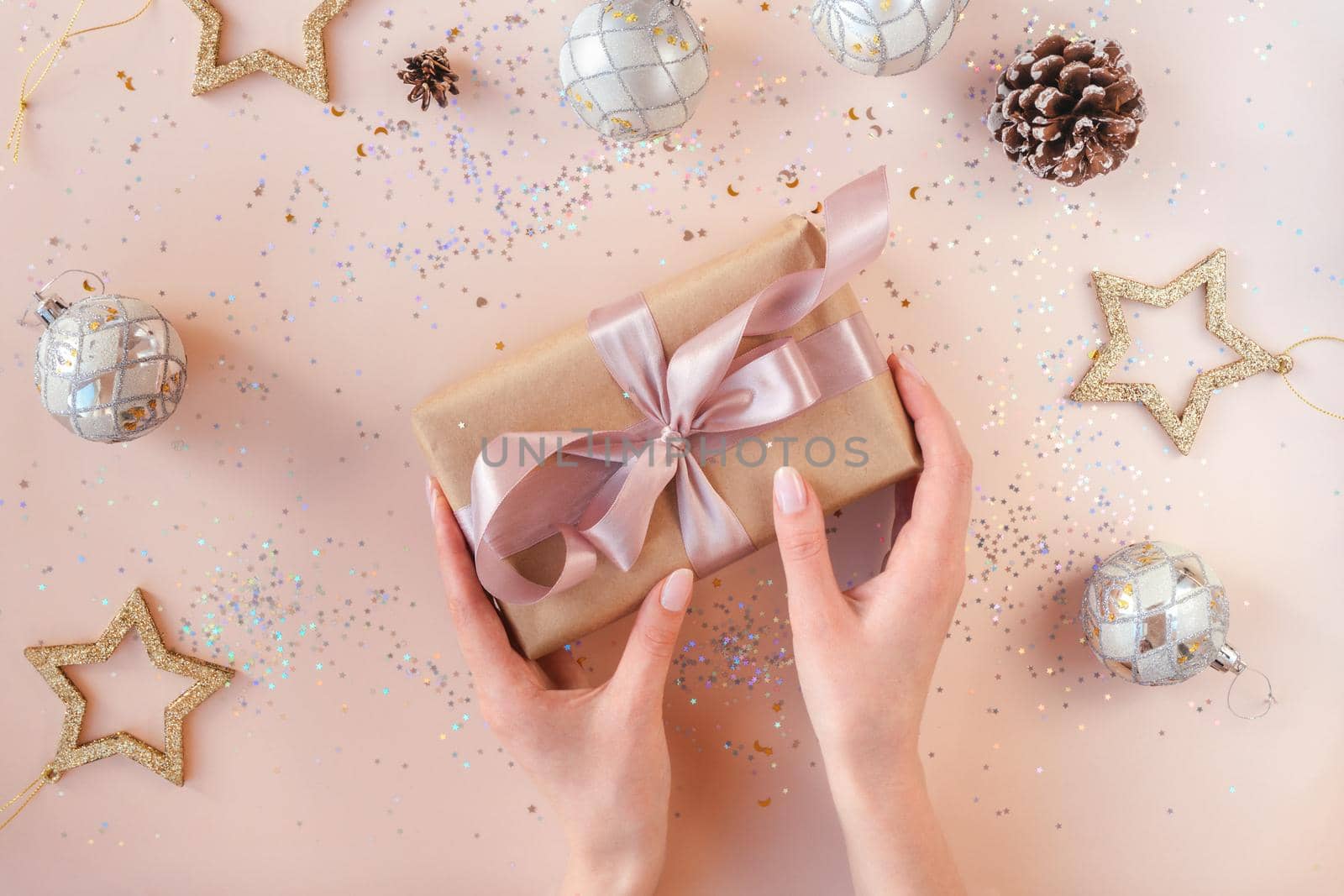 Female hands with natural manicure holding present in kraft paper with pink ribbon on soft beige background. Xmas composition. Flat lay. Happy holidays, New Year celebration and giving love concept.