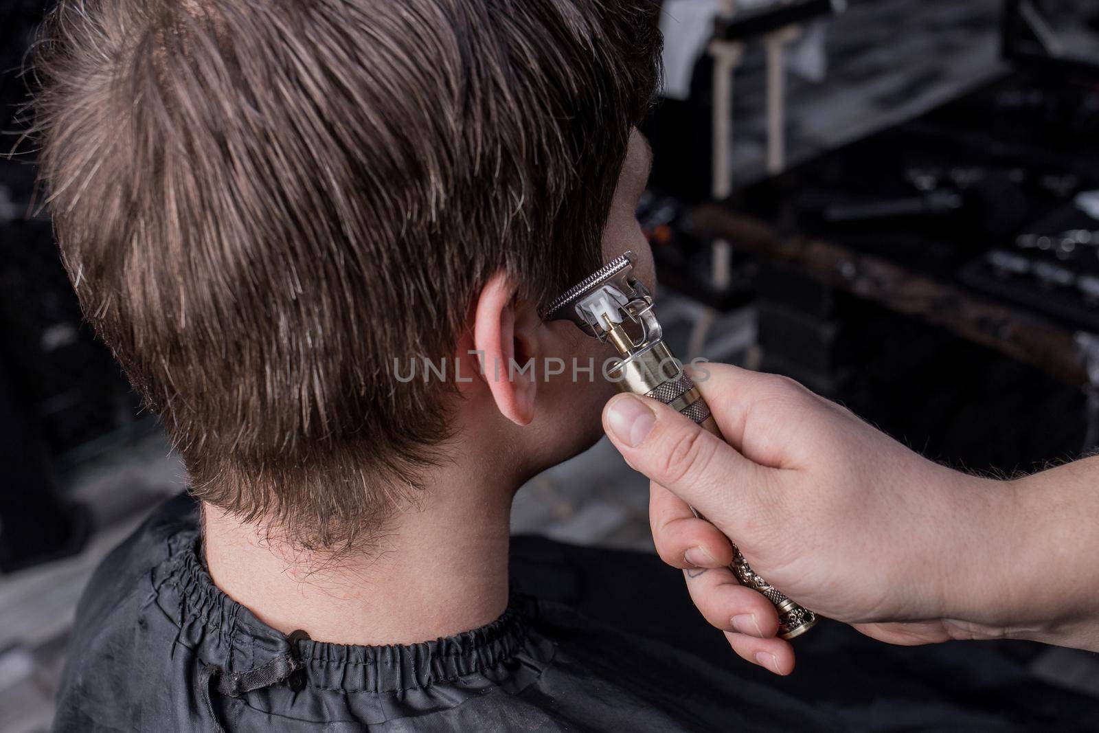 The hand of a barber or hairdresser cuts the dark long hair of the guy's client on his temple. Hairdressing.