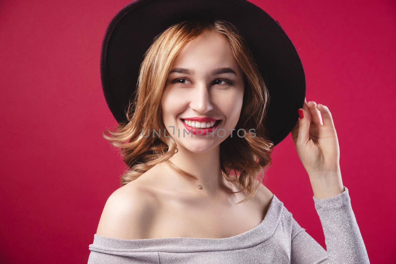 Fashionable attractive Caucasian girl in a large hat to large brim in a silver shiny dress on a red background. Studio retro portrait of a smiling young woman.