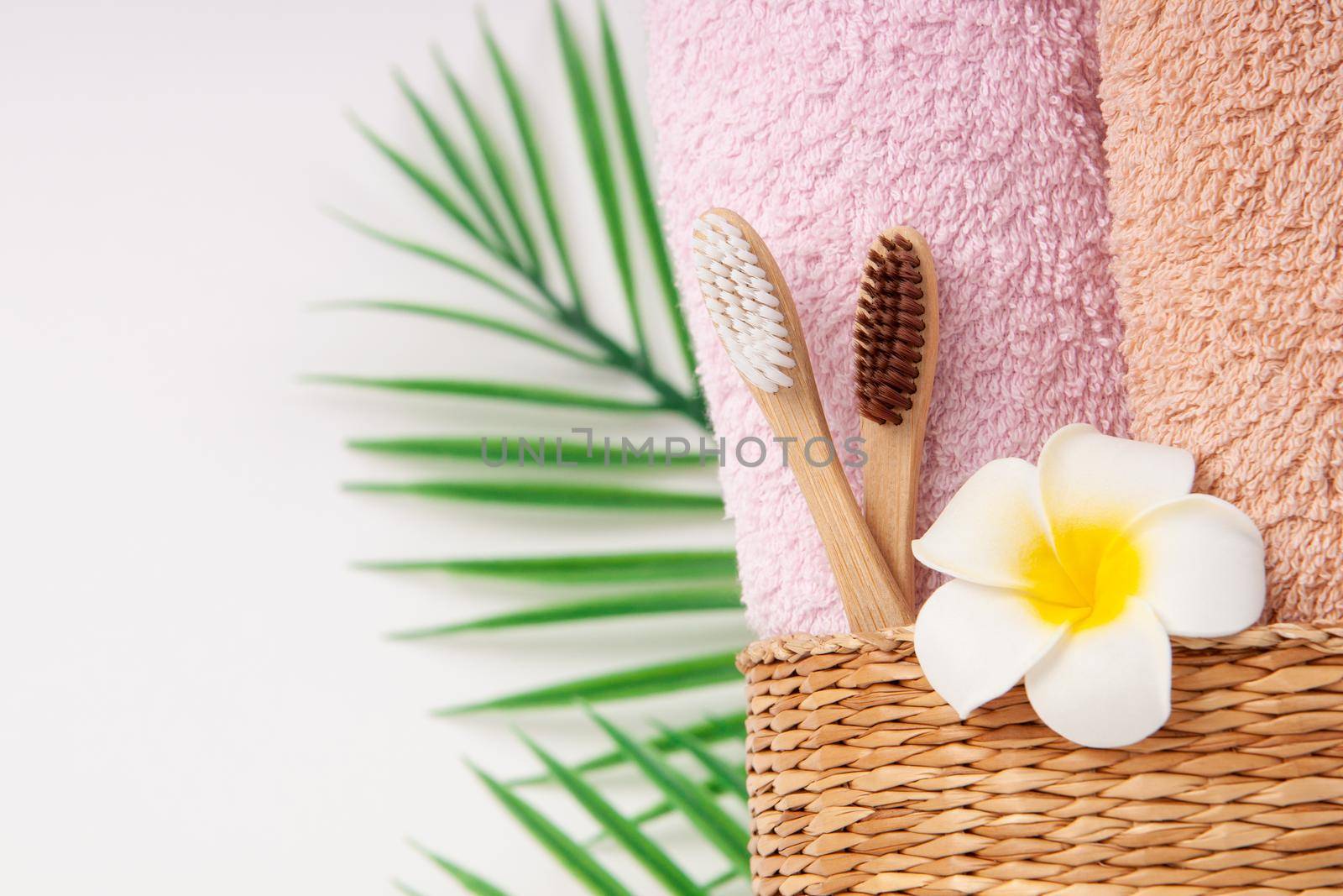 Bamboo toothbrushes with towels in a wicker wooden basket and palm leaves and plumeria flower with copy space on white background. Spa, healthy lifestyle and ecology concept. by chelmicky