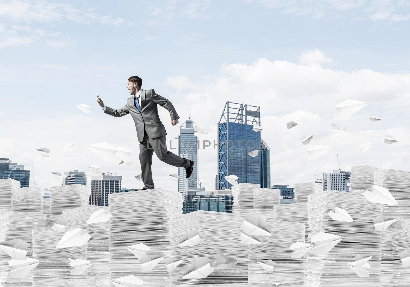 Businessman in black suit running with phone in hand among flying paper planes with cityscape on background. Mixed media.