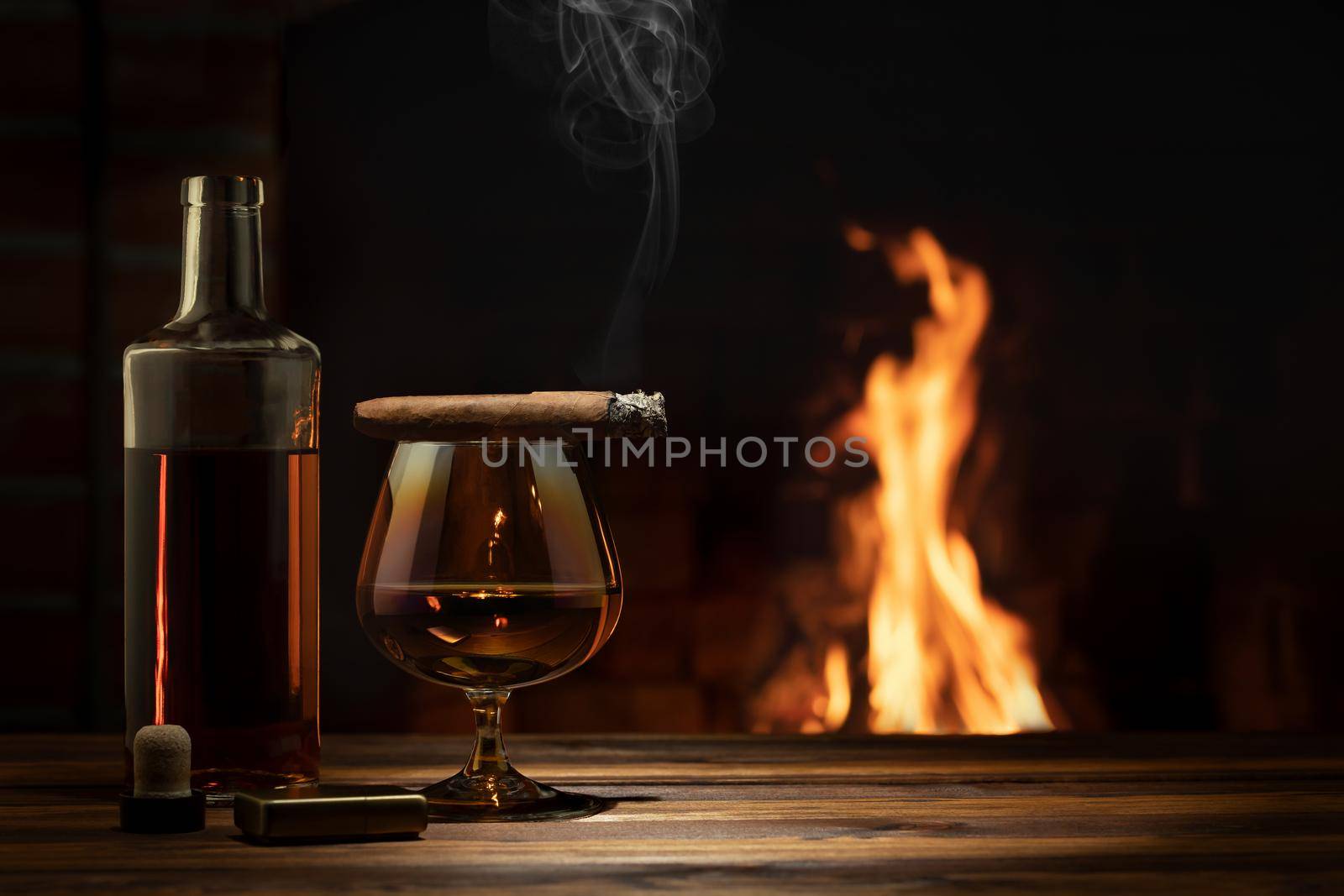 Glass of cognac, a cigar, a bottle on the table near the burning fireplace. Relaxation and enjoyment concept.
